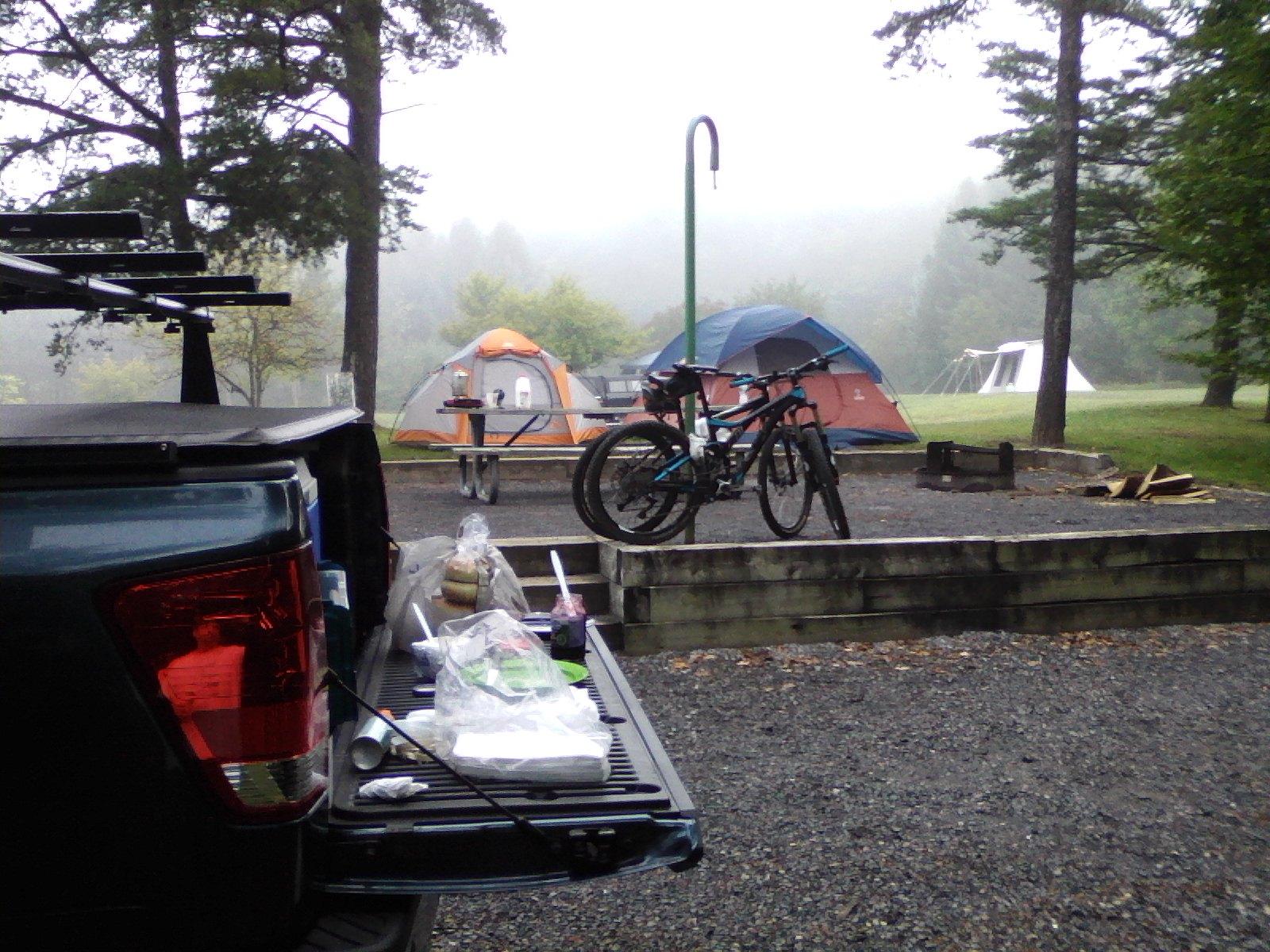 A view of a camping site featuring a pickup truck with an open tailgate displaying camping supplies, two bicycles parked nearby, and two colorful tents in the background. The scene is set in a foggy forested area, with trees surrounding the campsite. Allegrippis Trails mountain bike trail.