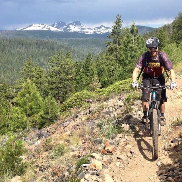 Canfield Brothers Yelli Screamy: A mountain biker navigating a rocky trail surrounded by lush greenery, with snow-capped mountains in the background under a partly cloudy sky.