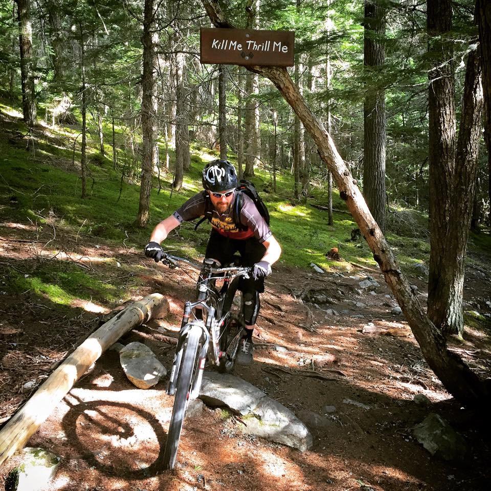 Niner R.I.P. 9 RDO: A mountain biker navigating a rocky trail in a forested area, with a wooden sign above reading "Kill Me Thrill Me." The scene features lush greenery, dirt paths, and scattered rocks. The biker is wearing a helmet and protective gear, showing intense focus as he maneuvers over a log on the trail.