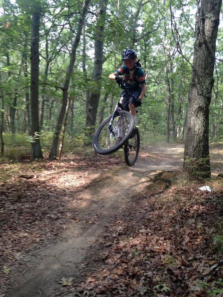 A young rider performing a jump on a mountain bike while navigating a dirt trail in a dense forest. The scene is filled with greenery, and fallen leaves cover the ground. The rider is wearing a helmet and biking gear, showcasing an action-packed moment in an outdoor setting.