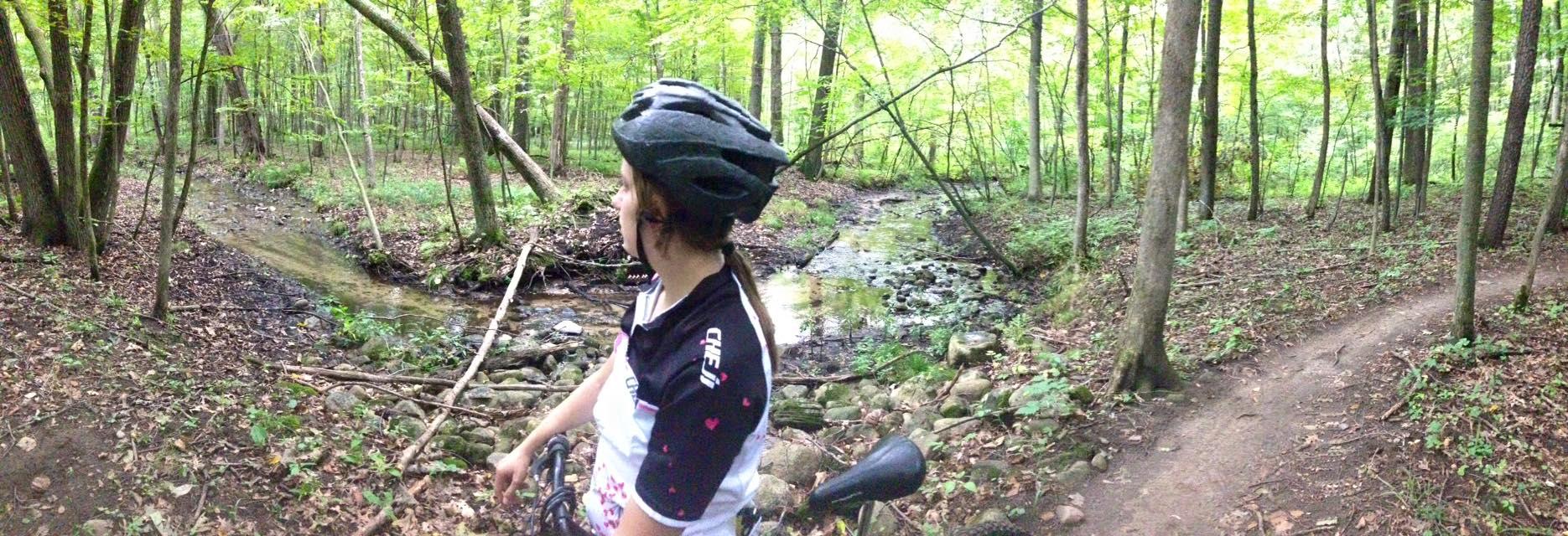 A cyclist wearing a helmet and a colorful jersey is resting beside a small stream in a lush green forest. The scene features tall trees and a winding dirt path, with fallen leaves and rocks along the water's edge. The cyclist is turned slightly, looking towards the stream and enjoying the serene natural surroundings. Luton Park Trail mountain bike trail.