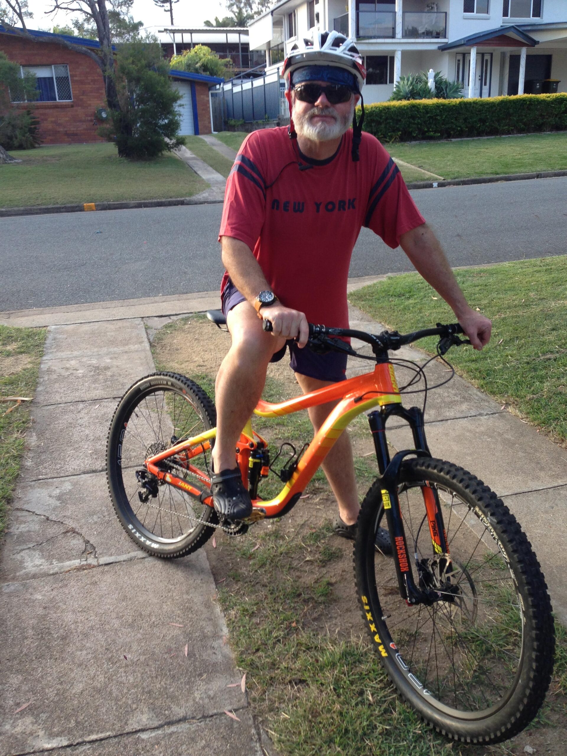 Giant Reign: An older man with a white beard is sitting on an orange and yellow mountain bike on a sidewalk. He is wearing a red "NEW YORK" t-shirt, black shorts, and a white bicycle helmet. The background features residential houses and a grassy area. The man is looking directly at the camera, ready to ride.