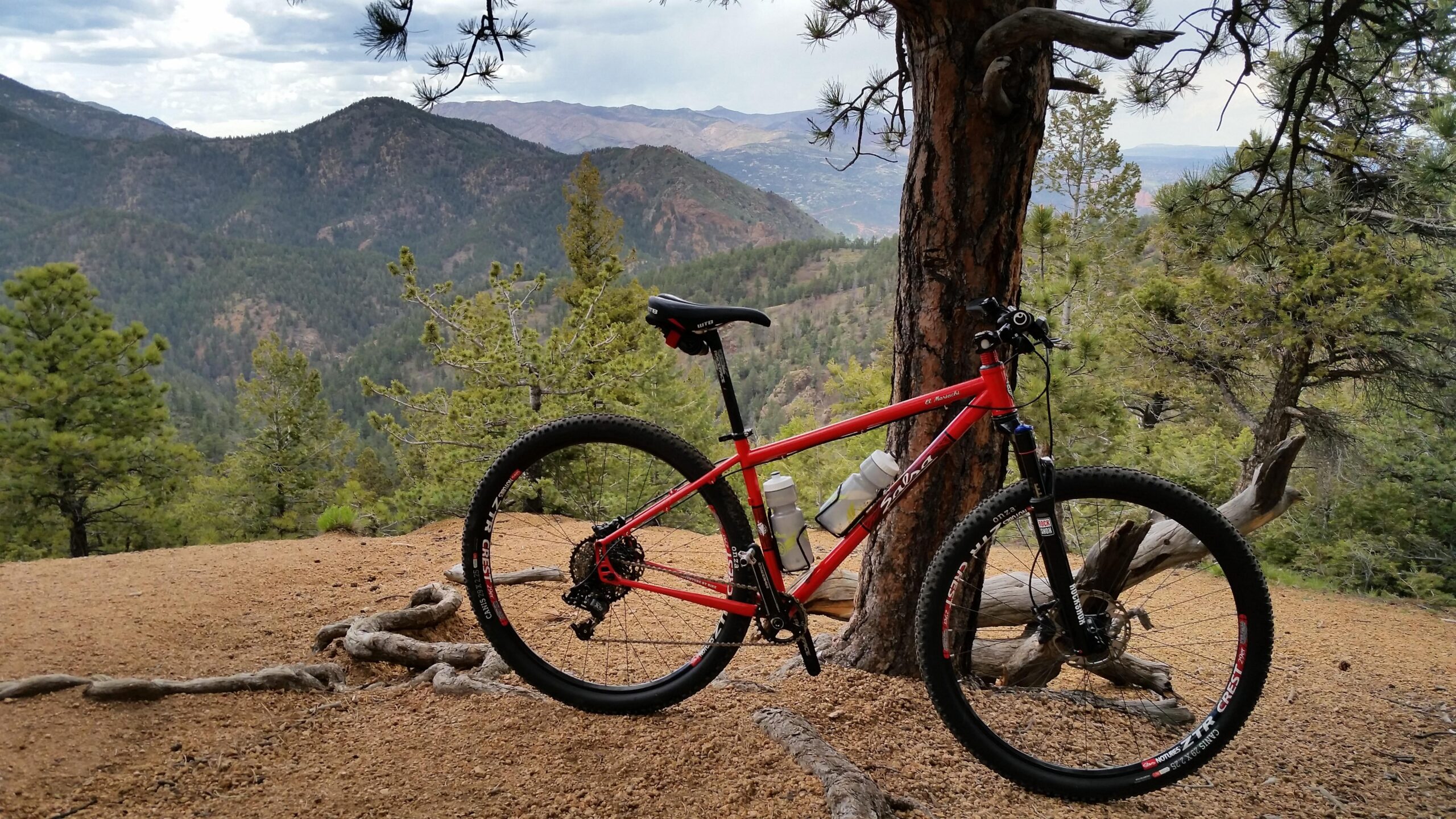 Salsa El Mariachi: A red mountain bike leaning against a tree on a dirt trail. In the background, a scenic view of green hills and mountains under a cloudy sky.