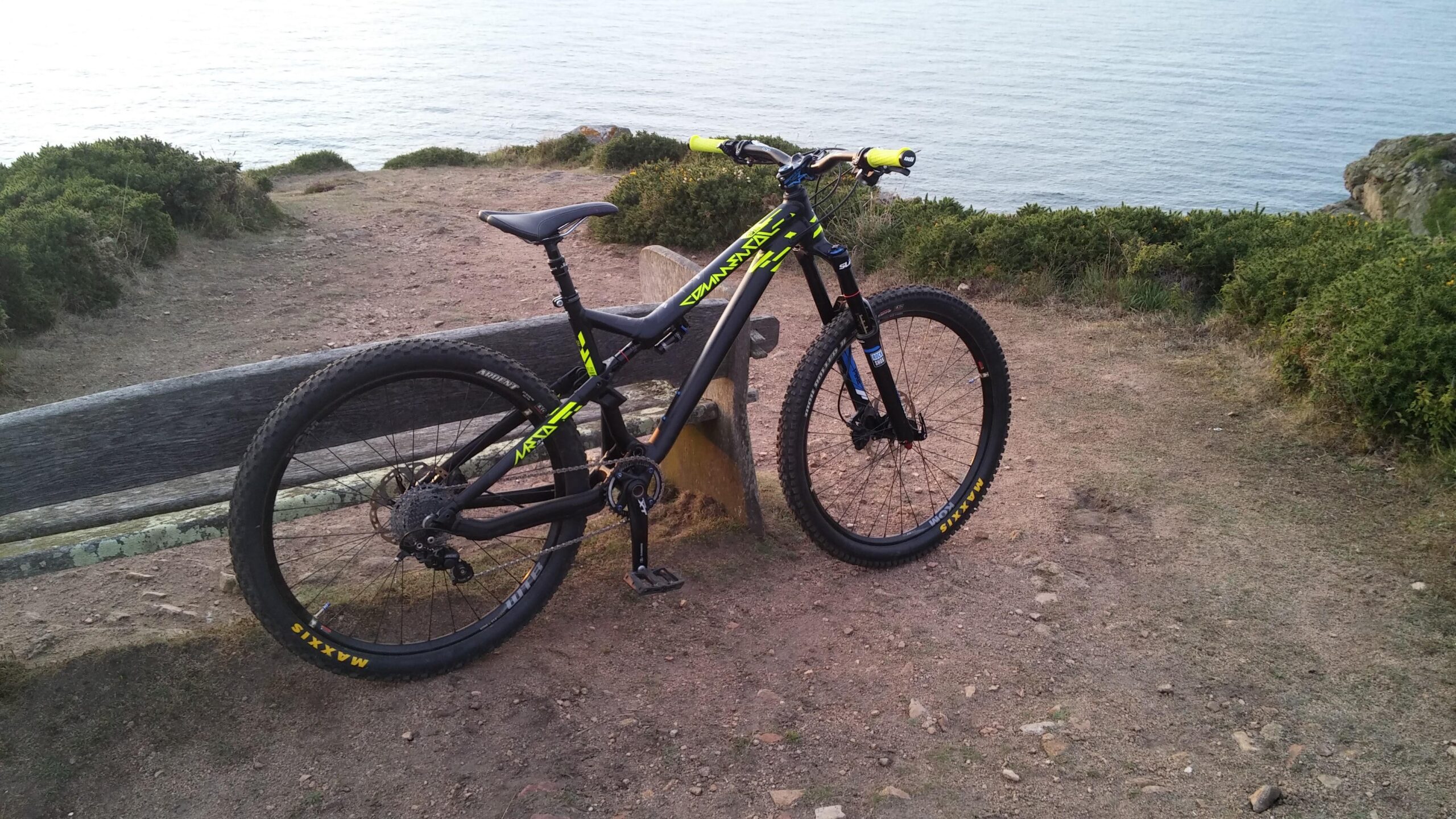 Commencal Meta AM V4: A mountain bike with a black frame and bright yellow accents is parked on a dirt trail near the edge of a cliff, overlooking a body of water. Surrounding the bike are grassy patches and shrubs. The scene is tranquil, with the sun reflecting off the water in the background.