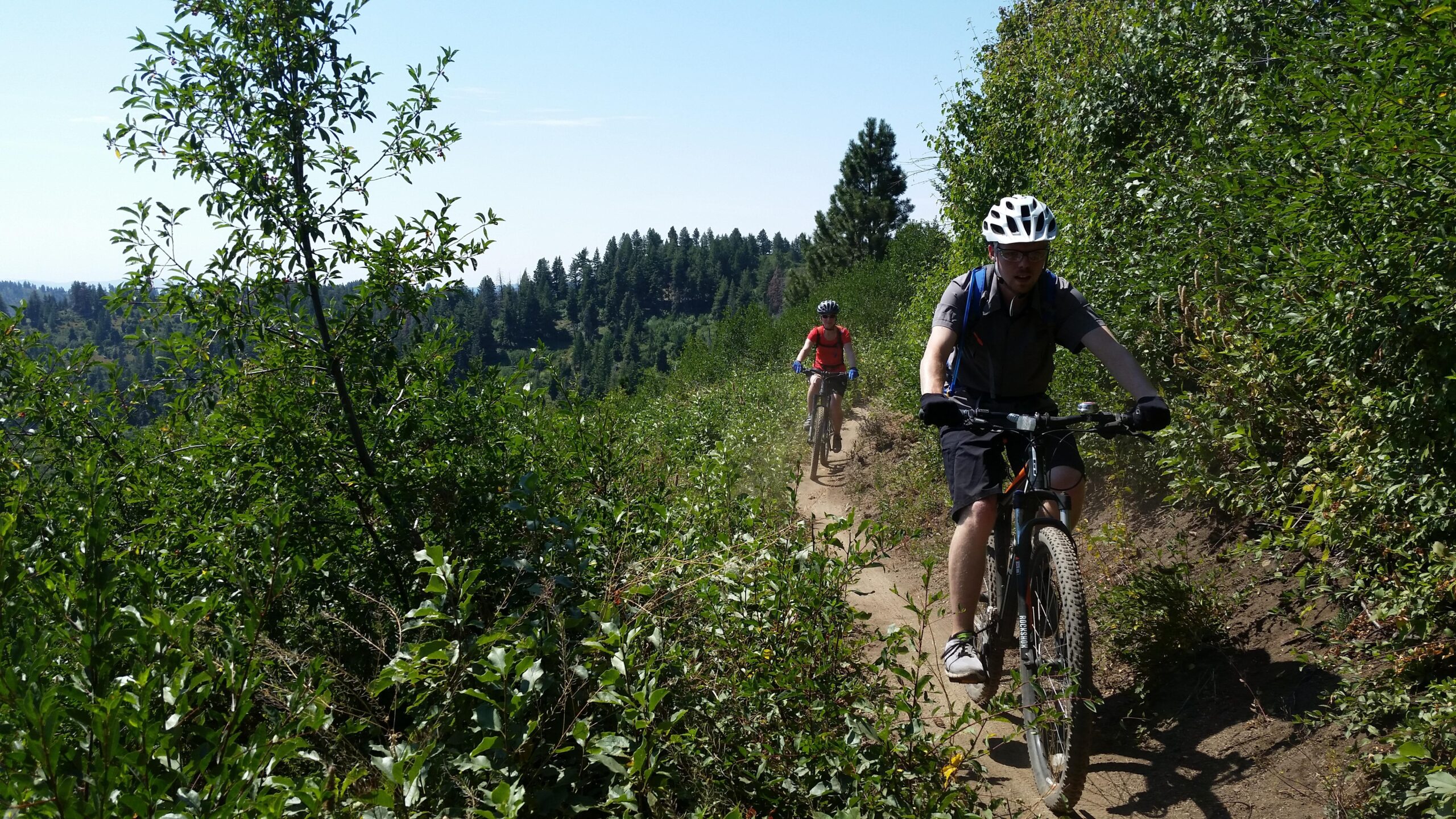 Two mountain bikers ride along a narrow dirt trail surrounded by greenery. The background features a scenic view of trees and hills under a clear blue sky. One biker is dressed in a red shirt, while the other wears a gray helmet and backpack. Around the Mountain mountain bike trail.