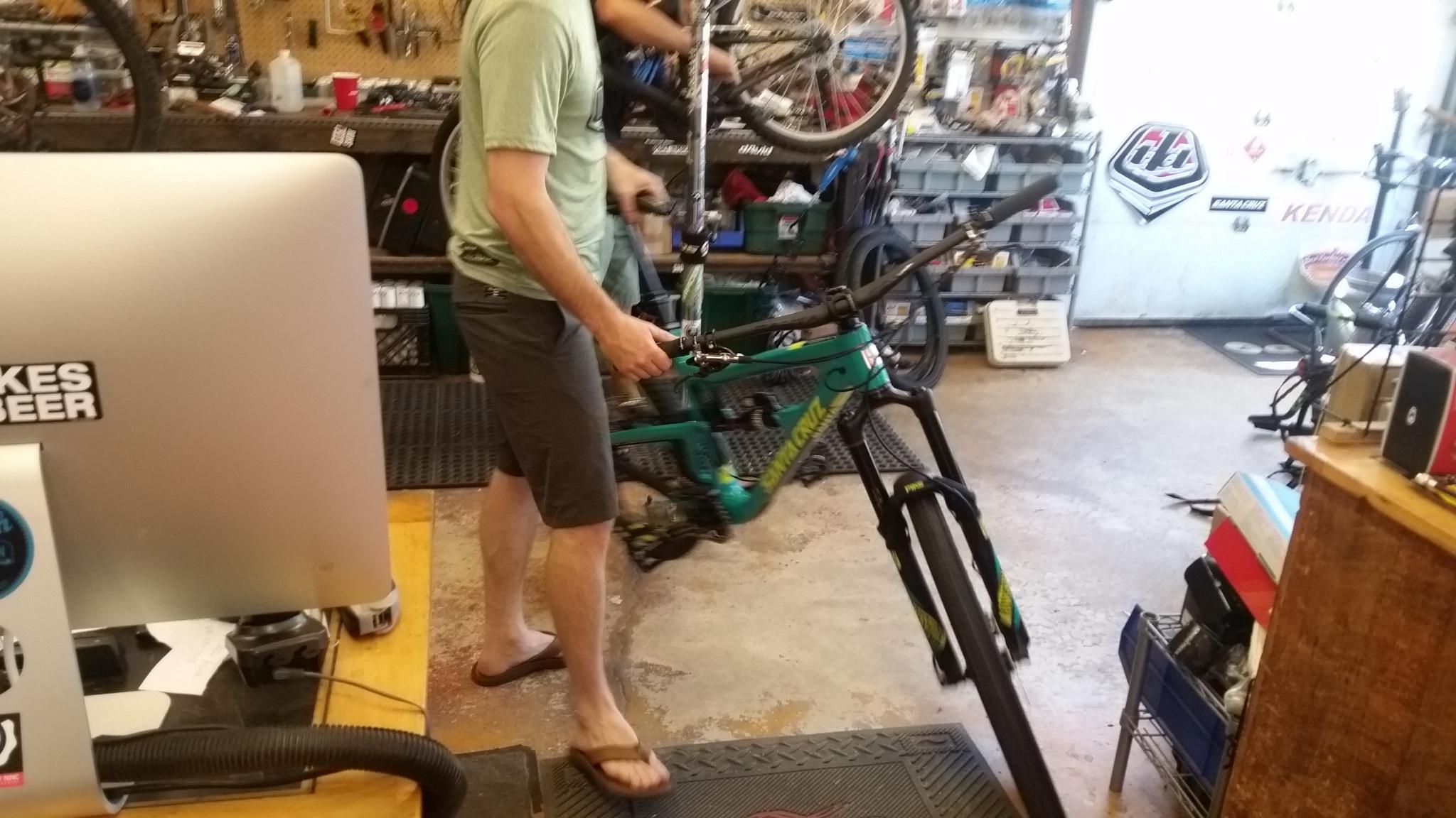 A person wearing a green shirt and shorts adjusts a mountain bike in a bustling bike shop filled with tools, bike parts, and repair stations. The bike being worked on is turquoise and prominently displayed in the foreground, while another bike is being lifted in the background. An Apple computer with a "Bikes Beer" sticker is visible on a workbench.