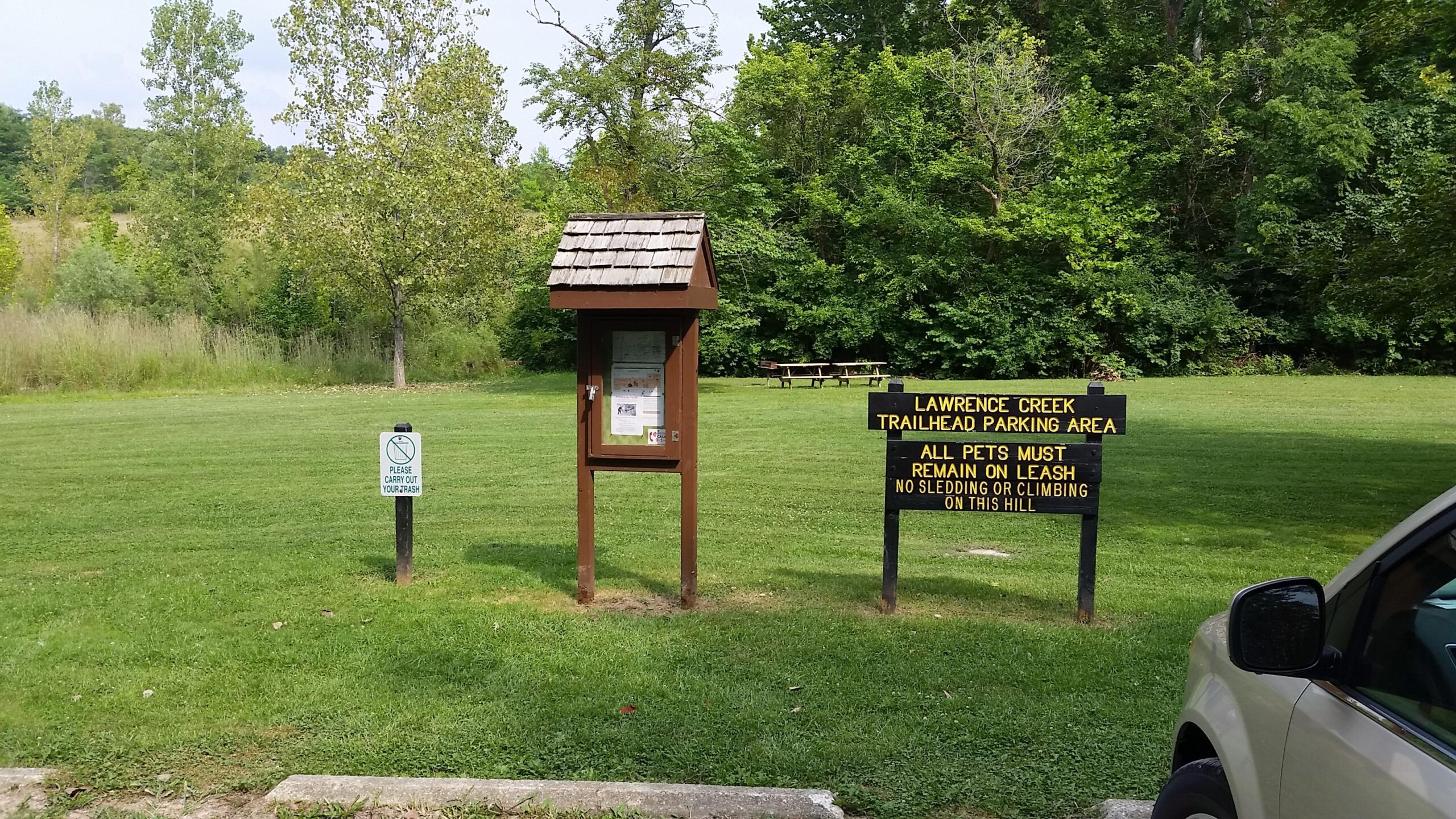 A grassy area with a sign indicating "Lawrence Creek Trailhead Parking Area" and rules about pets. There is a wooden information kiosk nearby, and a small sign that says "Please carry out your trash." Trees and a picnic area are visible in the background, with a part of a car in the foreground. Fort Harrison State Park mountain bike trail.