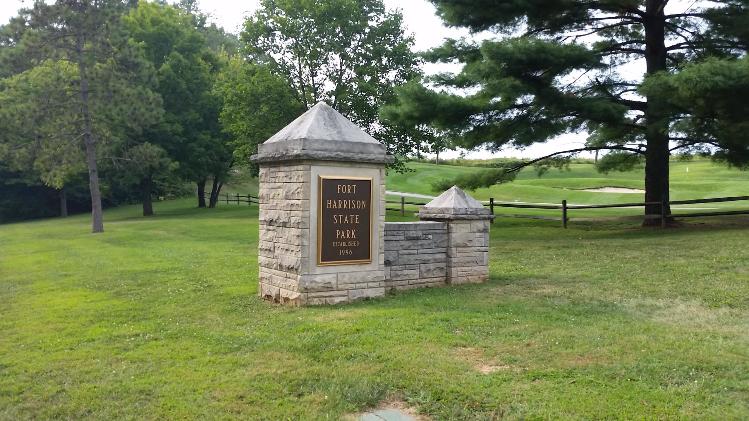 Sign marking the entrance to Fort Harrison State Park, with a stone structure, greenery, and a grassy area in the background. The sign displays the park's name and the year it was established, 1996. Fort Harrison State Park mountain bike trail.