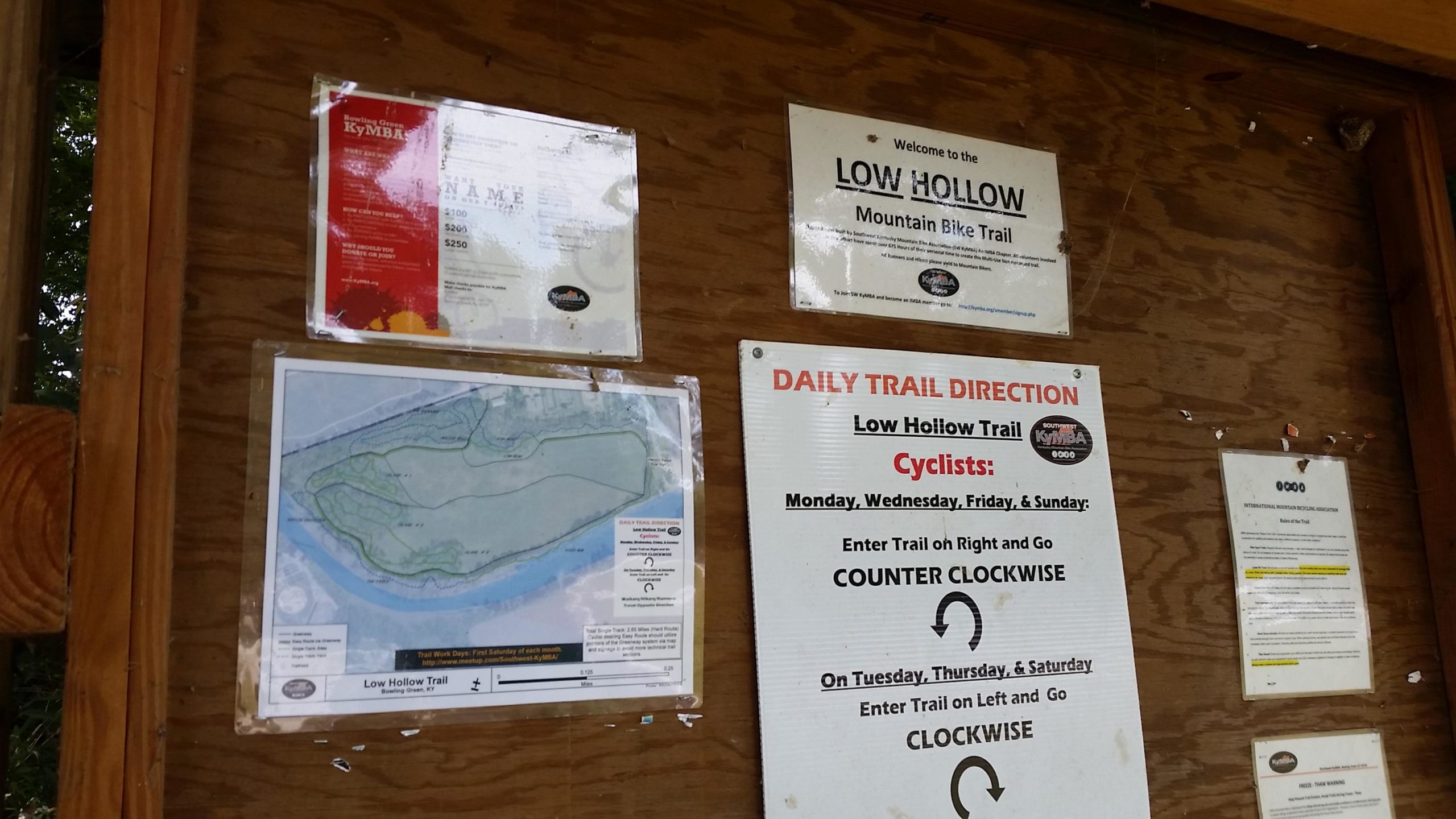 A wooden trail information board displaying various notices about the Low Hollow Mountain Bike Trail. It includes a map of the trail, daily trail directions for cyclists, and information about the Kymba organization. The board has postings about trail usage guidelines and volunteer opportunities. Low Hollow mountain bike trail.