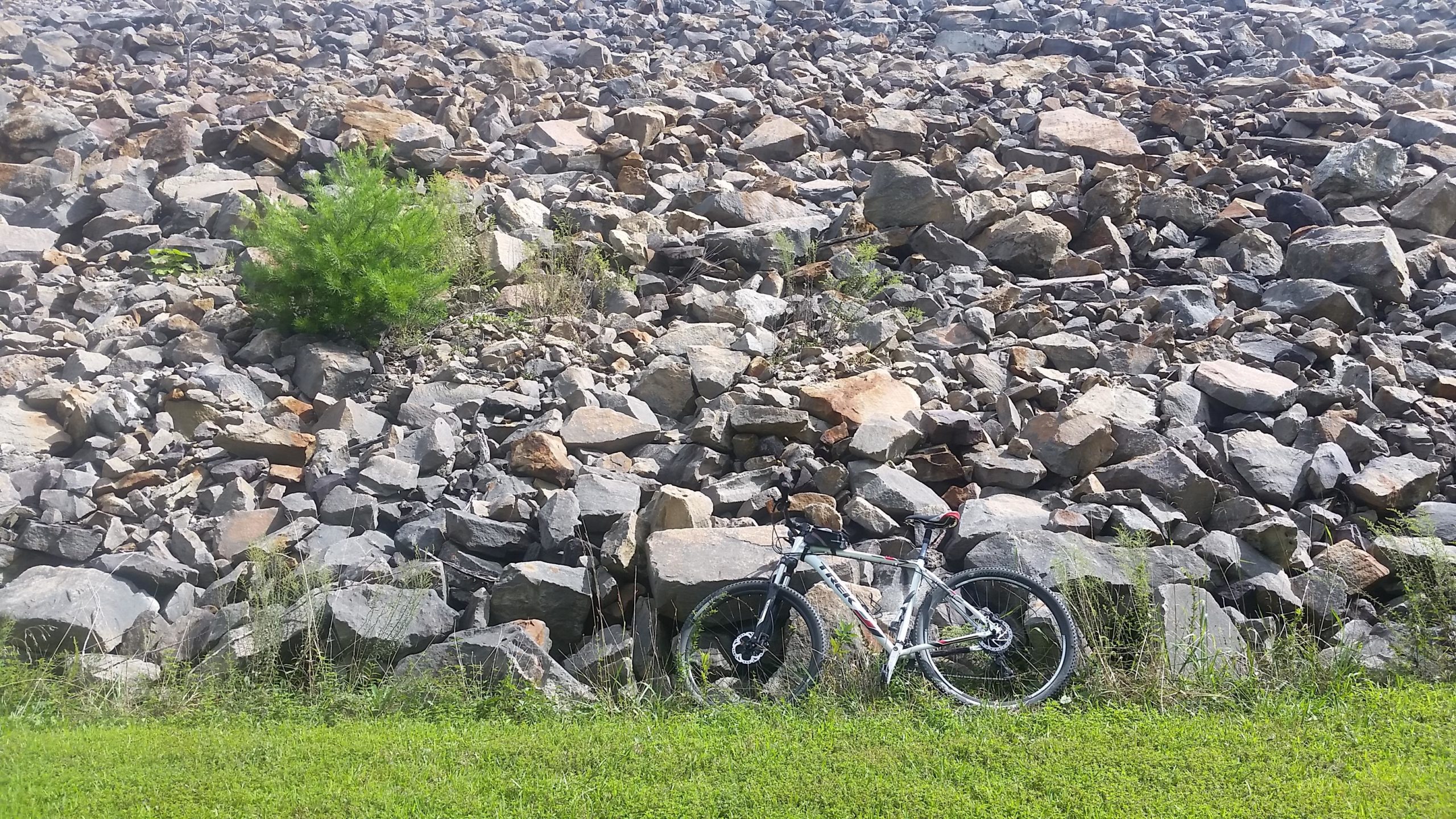 A mountain bike leaning against a rocky slope, with patches of green grass and small plants in the foreground. The background is filled with a diverse arrangement of large stones and boulders. Raccoon Mountain Trail Network mountain bike trail.