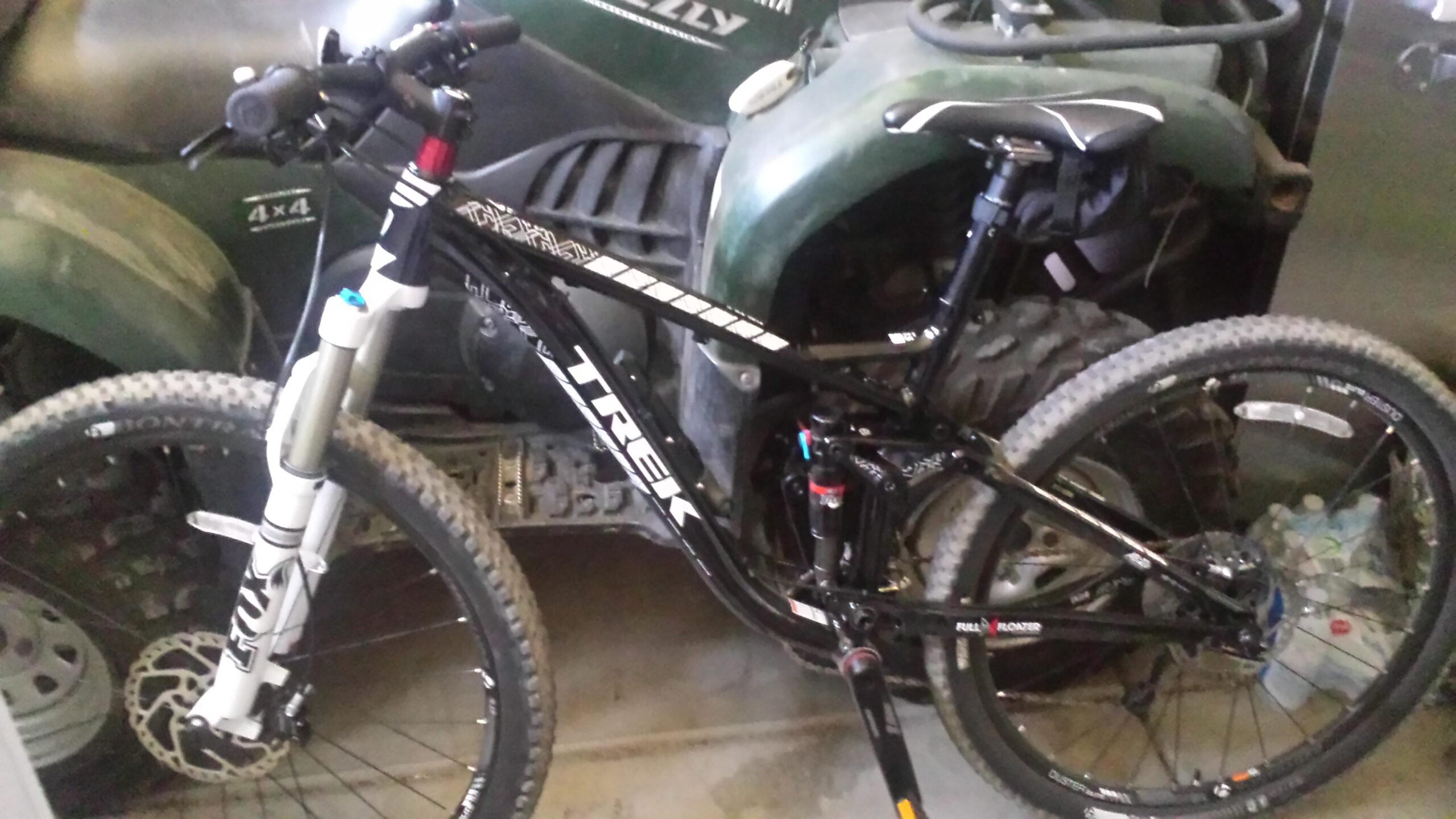 Trek EX 8: A black Trek mountain bike with a full suspension system, featuring large knobby tires and front disc brakes, is parked indoors next to a green all-terrain vehicle (ATV). The bike has a sleek design, with white branding and visible suspension components.