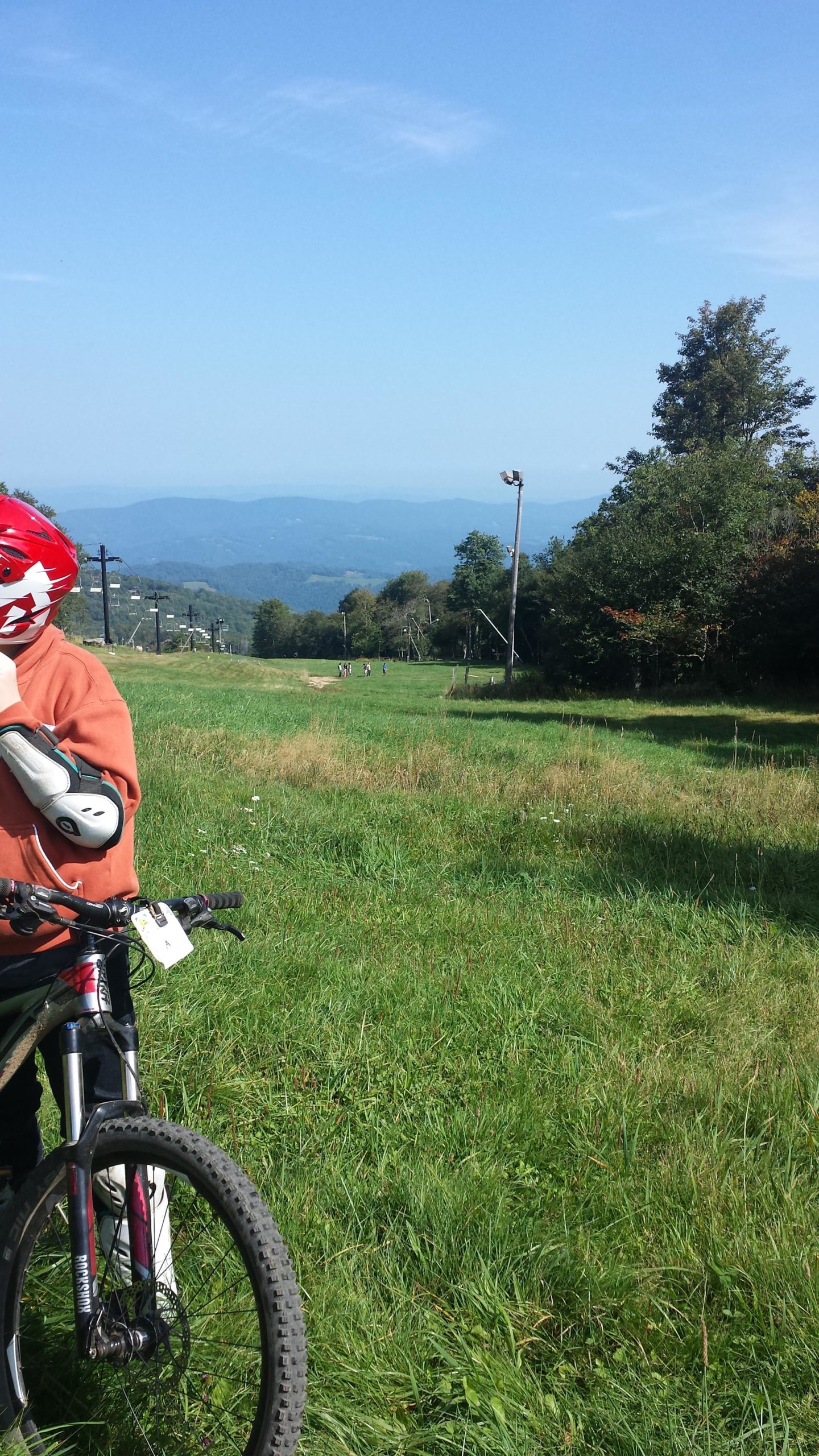 A person wearing a red helmet and protective gear stands next to a mountain bike on a grassy slope. In the background, a scenic view of rolling hills and mountains is visible under a clear blue sky. People can be seen in the distance, enjoying outdoor activities. Emerald Outback mountain bike trail.