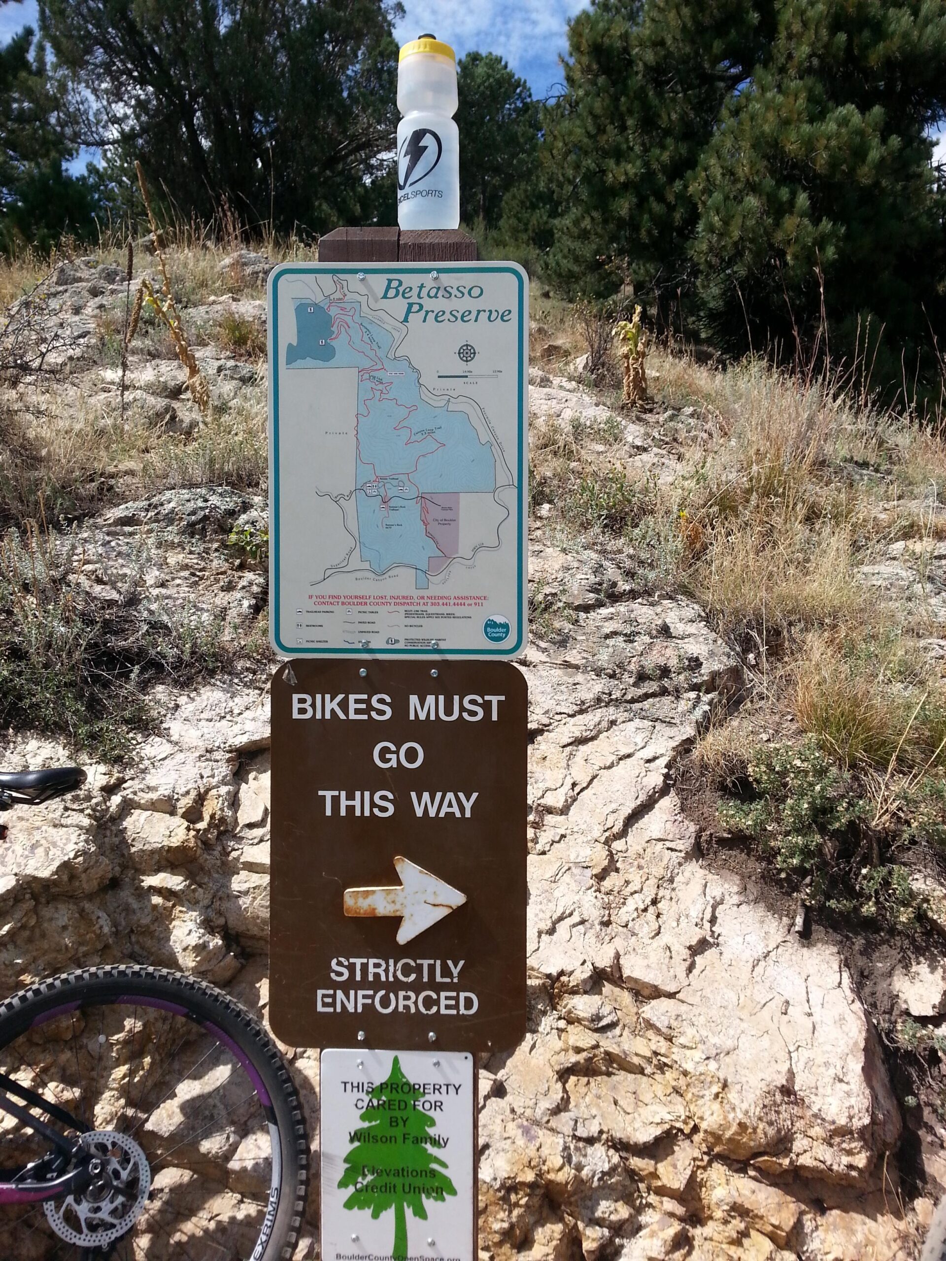 Signage at Betasso Preserve detailing a map of the area, indicating the direction for bicycles with a "Bikes Must Go This Way" arrow. A water bottle is resting on top of the map sign. The background features rocky terrain and pine trees under a partly cloudy sky. Betasso Preserve mountain bike trail.