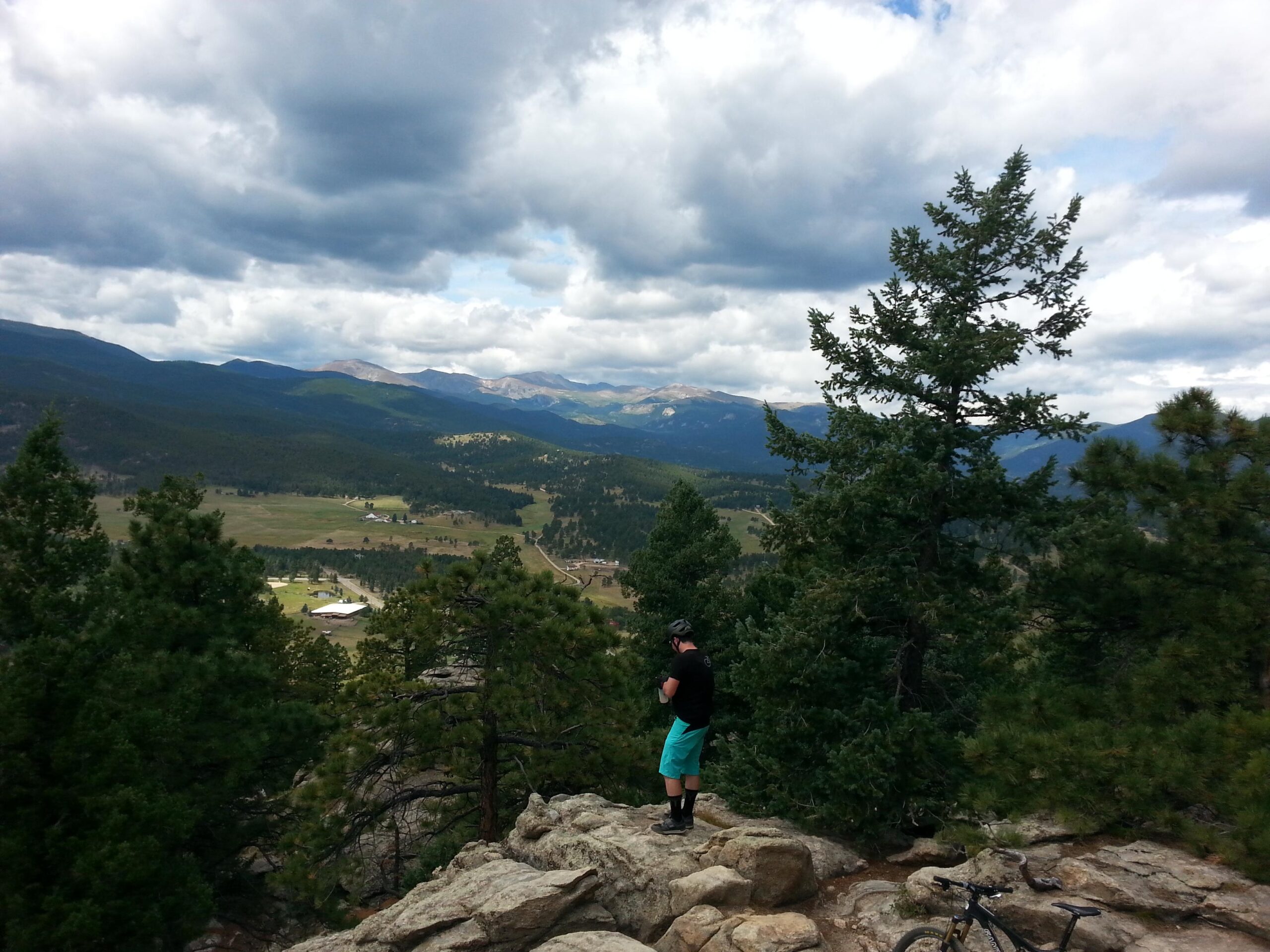 A person standing on a rocky outcrop overlooking a mountainous landscape, surrounded by pine trees and a cloudy sky. A bicycle is visible nearby. The view includes rolling hills and distant mountains. 3 Sisters / Alderfer mountain bike trail.