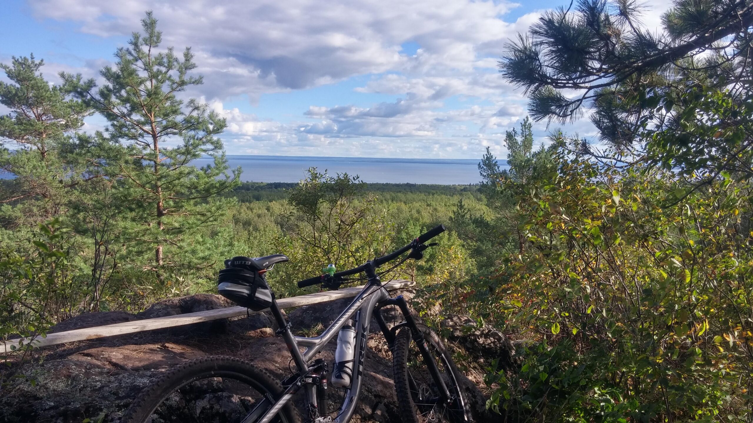A mountain bike rests on a rock ledge, surrounded by lush greenery and trees, overlooking a serene lake under a partly cloudy sky. Lester Park mountain bike trail.