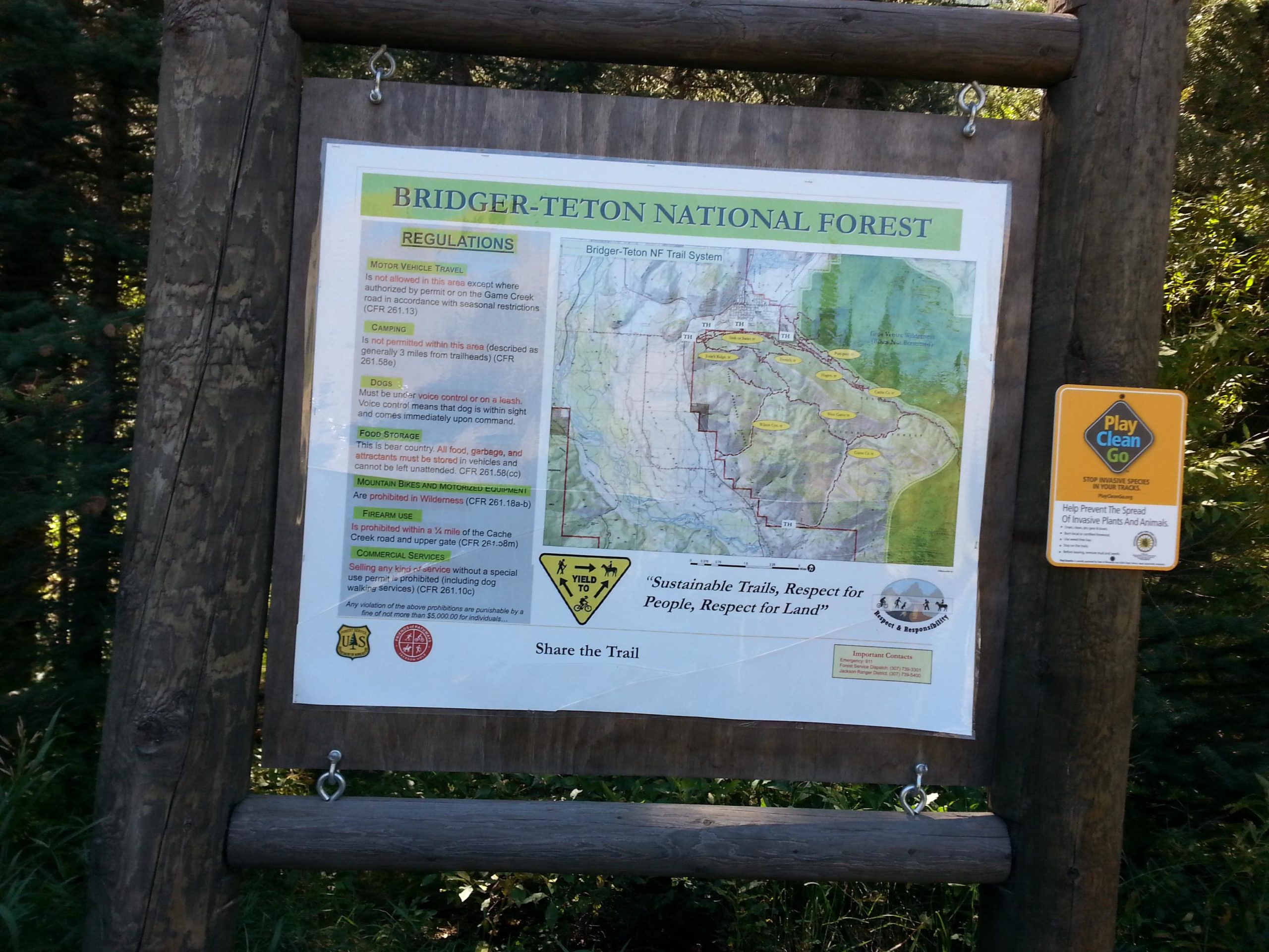 A wooden sign displaying a map of Bridger-Teton National Forest along with regulations for visitors. The map highlights trails and important locations within the forest, while the regulations include guidelines for motor vehicle travel, camping, dog control, food storage, and fire restrictions. Additionally, a "Play Clean Go" sign is attached, emphasizing the importance of preventing the spread of invasive plants and animals. Snow King Mountain mountain bike trail.