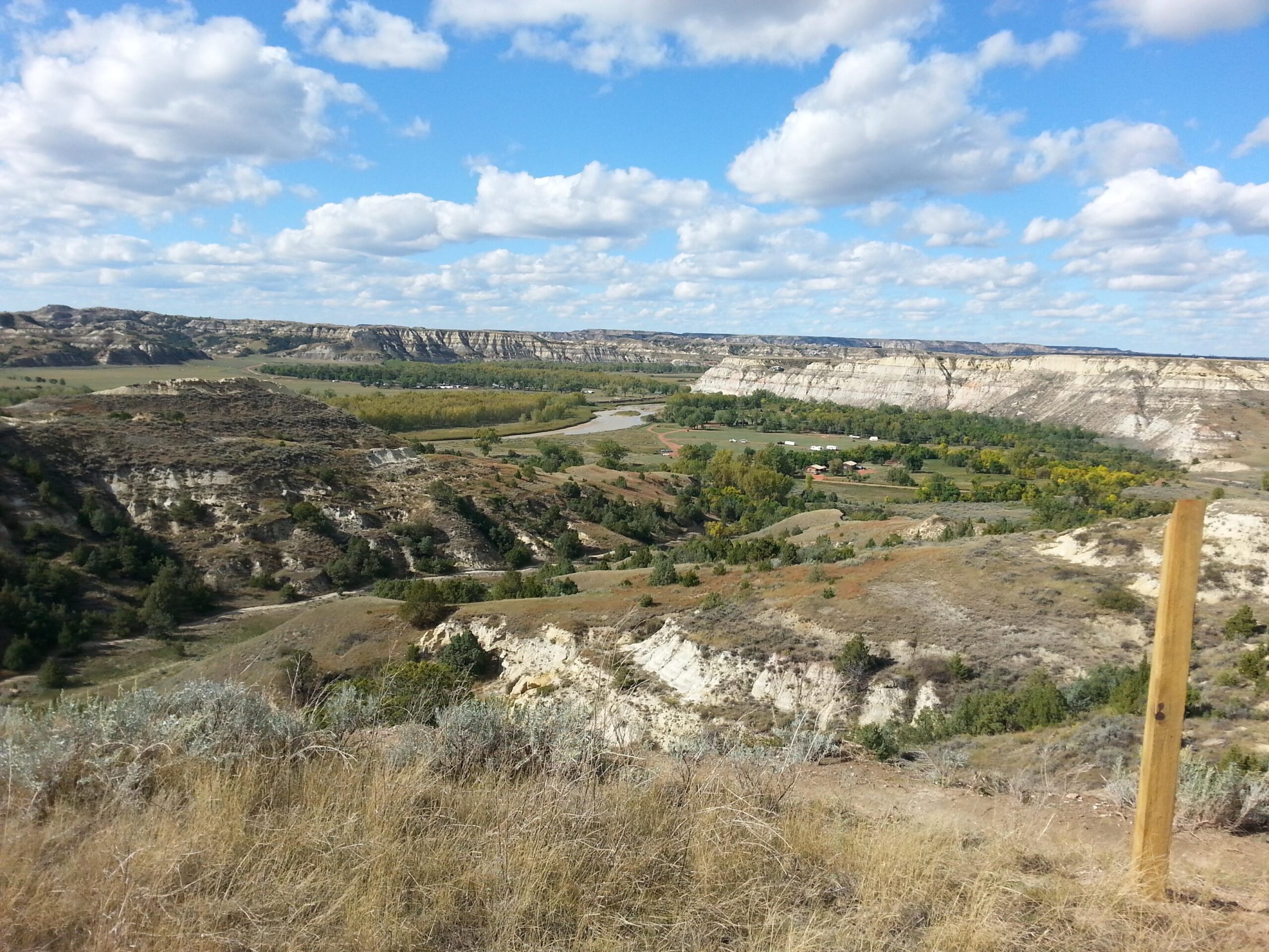 A panoramic view of a rugged landscape featuring hills and cliffs, with a winding river and lush green trees in a valley below. The sky is partly cloudy, adding depth to the scenic beauty of the area. In the foreground, dry grass and shrubs are visible, while a wooden post stands to the side. Maah Daah Hey mountain bike trail.