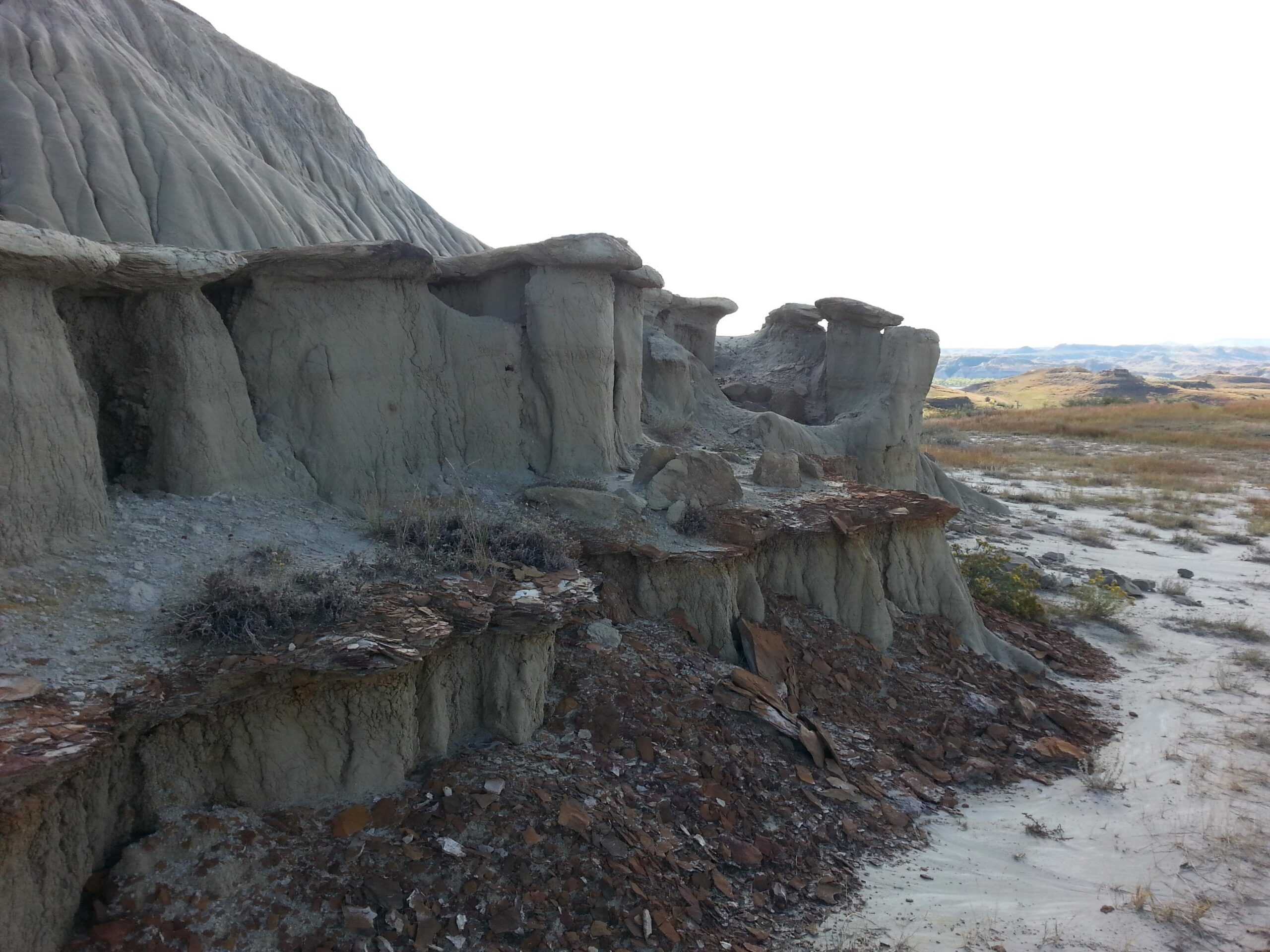 A rugged landscape featuring eroded rock formations with flat-topped mesas and layered sediment, set against a backdrop of rolling hills. Sparse vegetation is visible along the rocky terrain, showcasing natural geological structures under a bright sky. Maah Daah Hey mountain bike trail.
