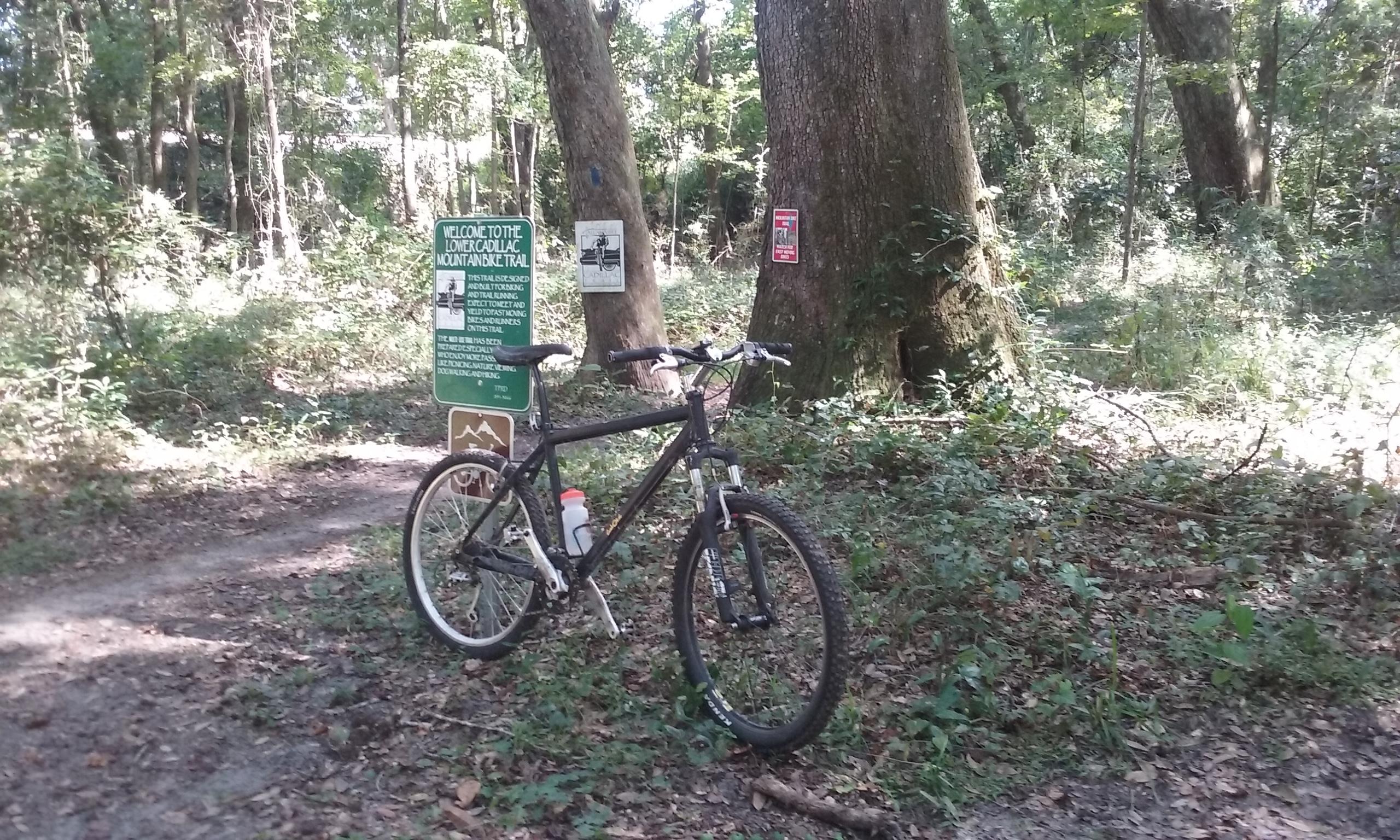 A mountain bike parked on a dirt trail in a wooded area, with a green sign in the background that welcomes visitors to the Lower Cadallic Mountainside Trail. The scene features lush greenery and large trees, indicating a natural outdoor setting. Tom Brown / Lafayette Heritage Park mountain bike trail.