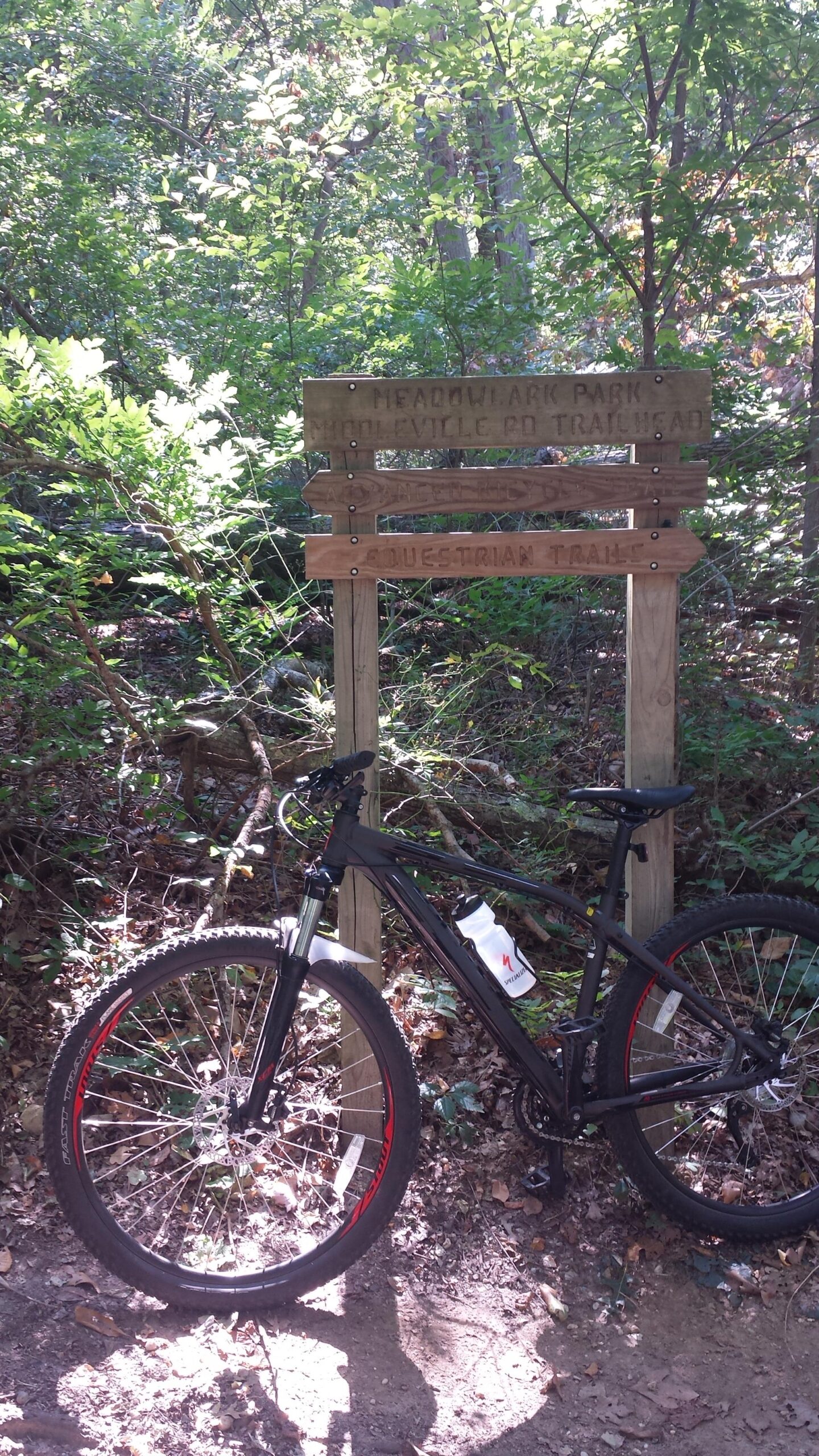 A mountain bike is parked next to a wooden trail sign in a lush green forest. The sign indicates the trailhead for Meadowlark Park and features directions for equestrian trails. Sunlight filters through the trees, highlighting the natural surroundings. Meadowlark Park mountain bike trail.