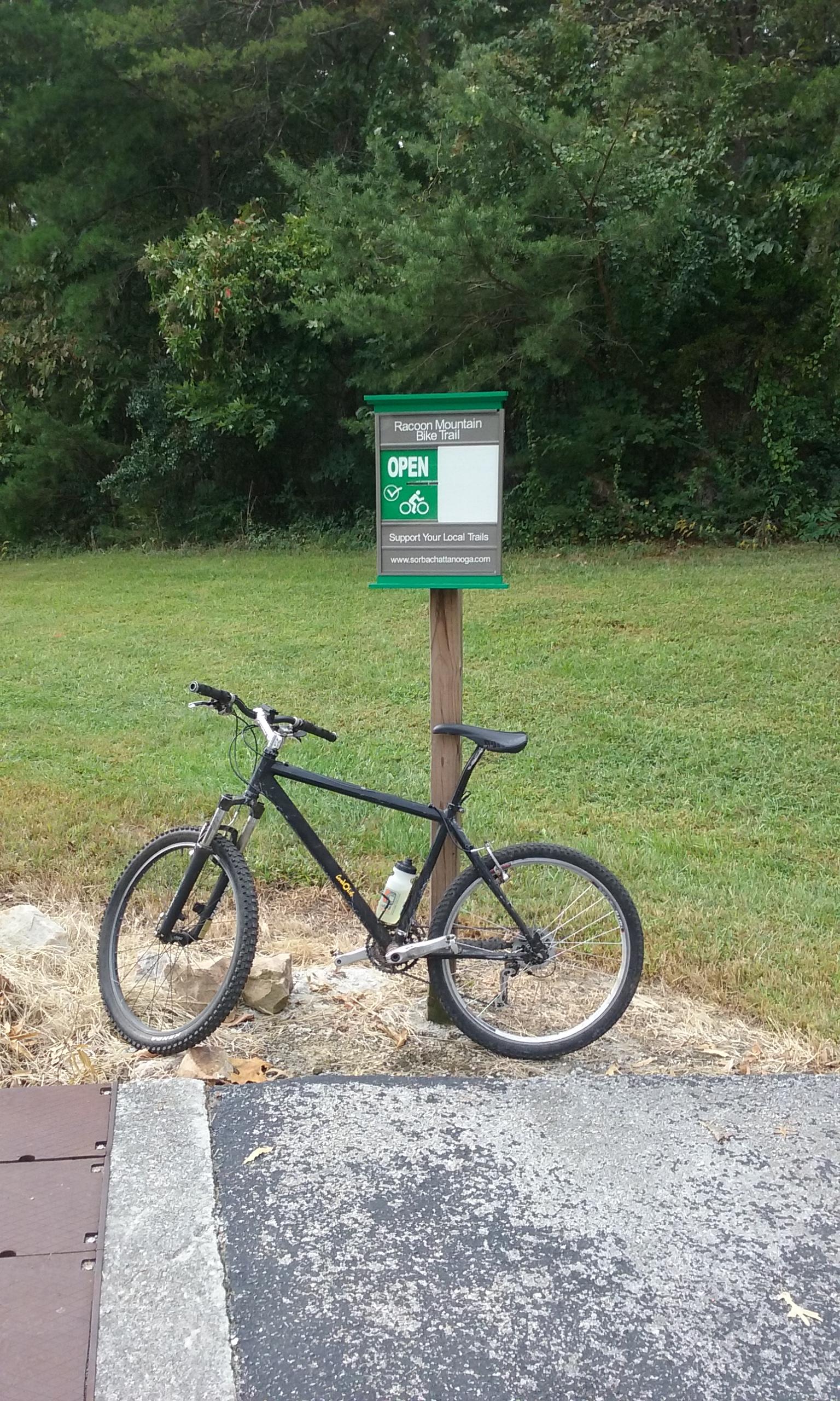 A black mountain bike parked beside a sign indicating the Raccoon Mountain Bike Trail is open, surrounded by green grass and trees in the background. The sign reads "Support Your Local Trails" and features a bicycle icon. Raccoon Mountain Trail Network mountain bike trail.