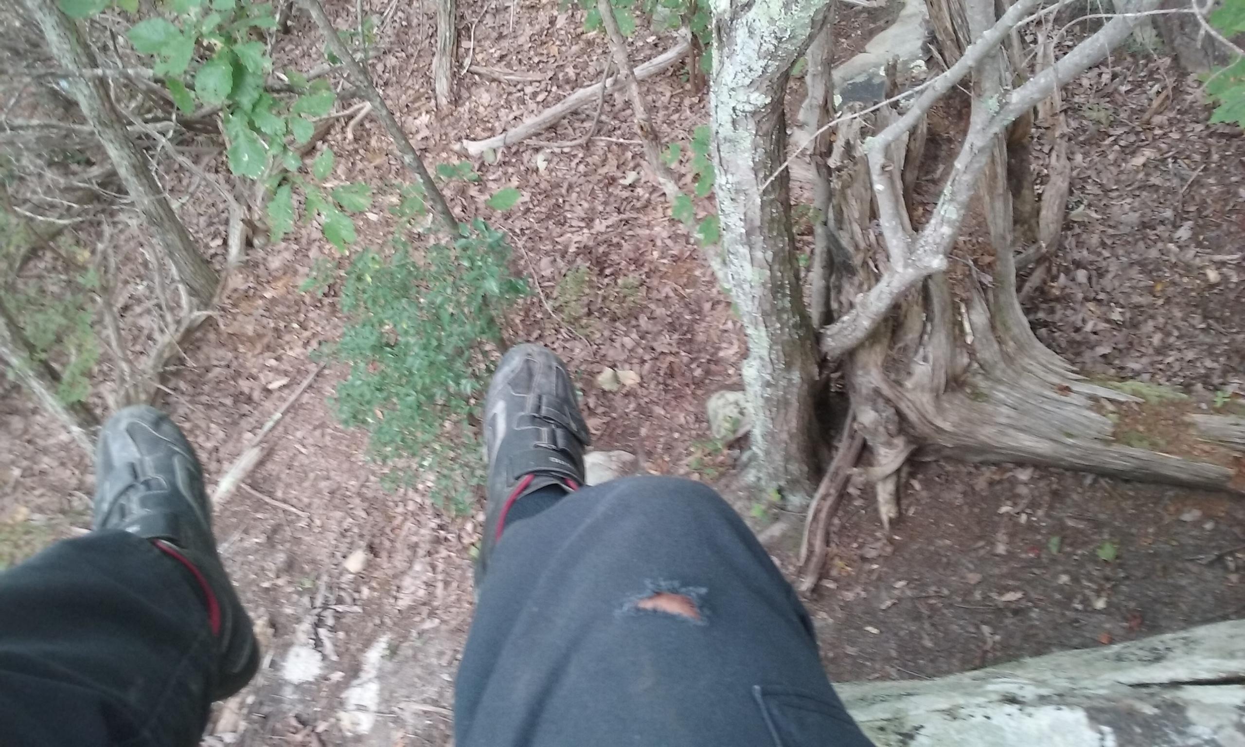 A close-up view of a person's legs and feet, hanging over the edge of a rocky ledge in a wooded area. The ground below is covered with leaves and small plants. The person is wearing a black pant with a visible tear and sturdy shoes. Surrounding them are trees and branches, creating a natural forest setting. Raccoon Mountain Trail Network mountain bike trail.