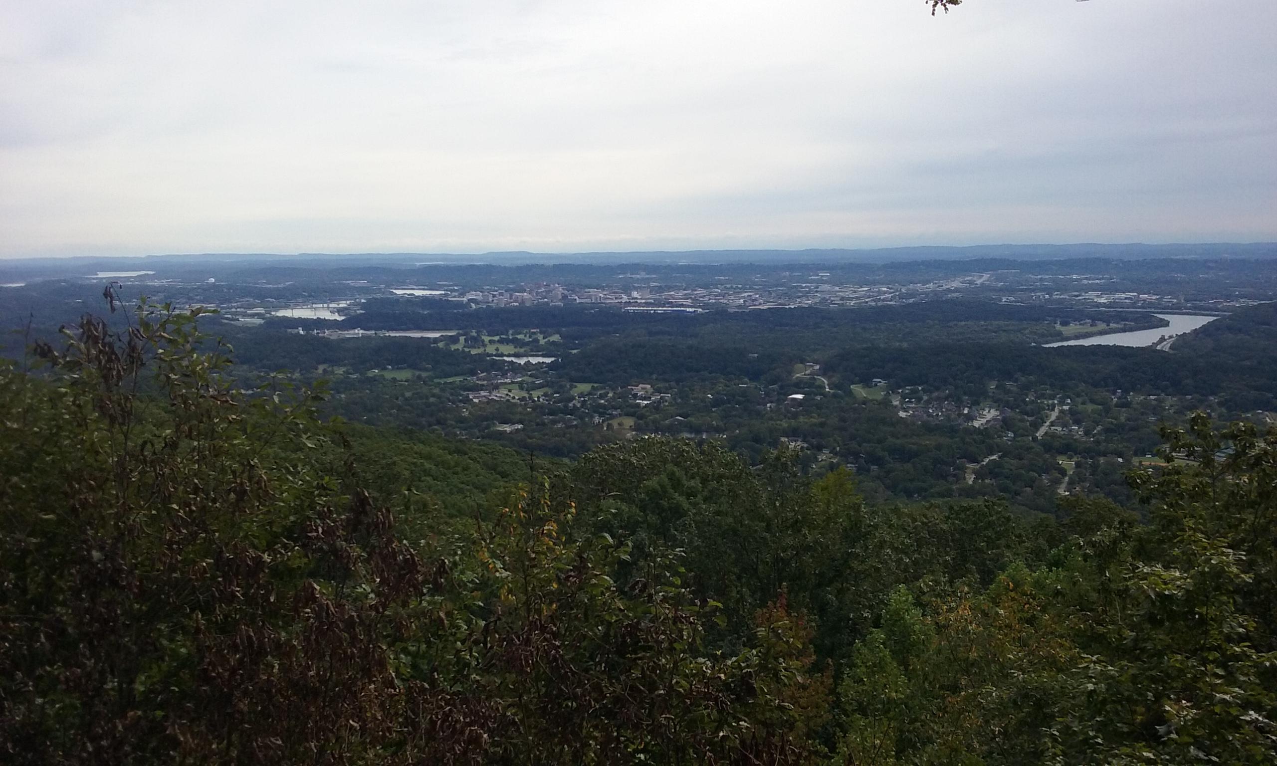 A scenic view from a mountain overlooking a lush green valley, dotted with patches of trees and small buildings. In the distance, a river meanders through the landscape, and a city skyline can be seen under a cloudy sky. Raccoon Mountain Trail Network mountain bike trail.