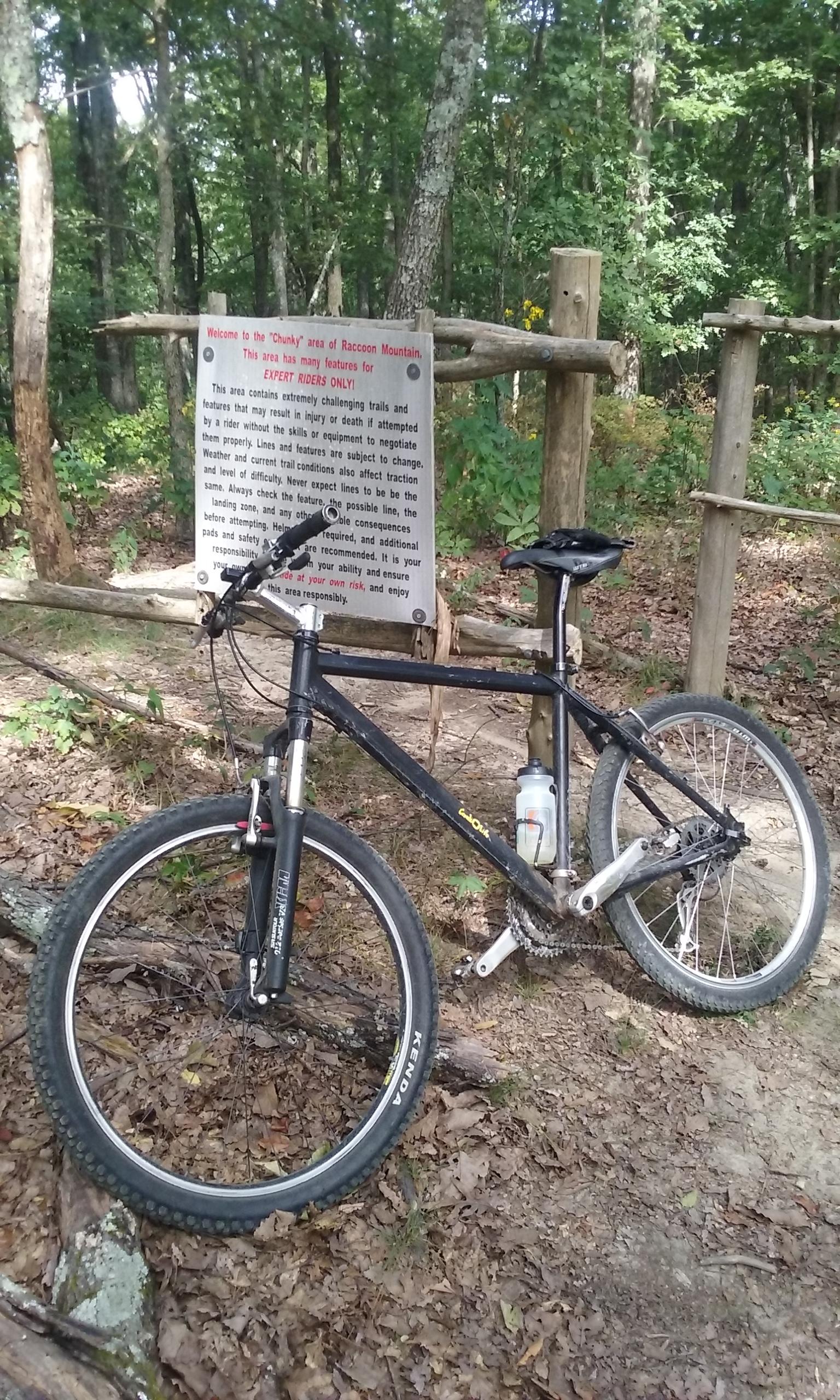 A black mountain bike is leaning against a wooden trail sign in a wooded area. The sign provides information about the "Chunky" area of Raccoon Mountain, warning that the trails are intended for expert riders only due to their challenging nature. Surrounding foliage includes trees and fallen leaves, indicating a natural outdoor environment. Raccoon Mountain Trail Network mountain bike trail.