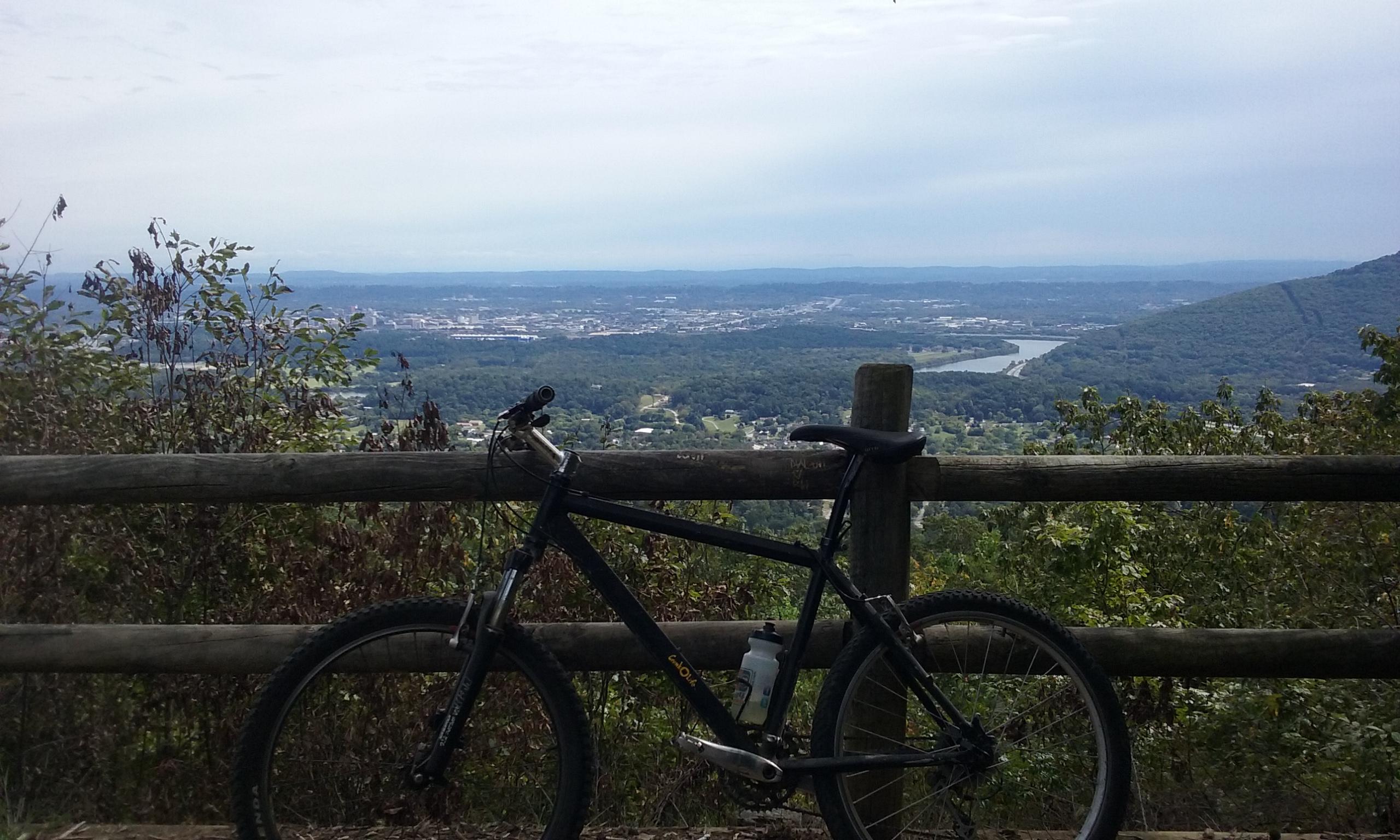 A mountain bike parked near a wooden railing, overlooking a scenic view of a valley with a river and distant mountains under a cloudy sky. Raccoon Mountain Trail Network mountain bike trail.