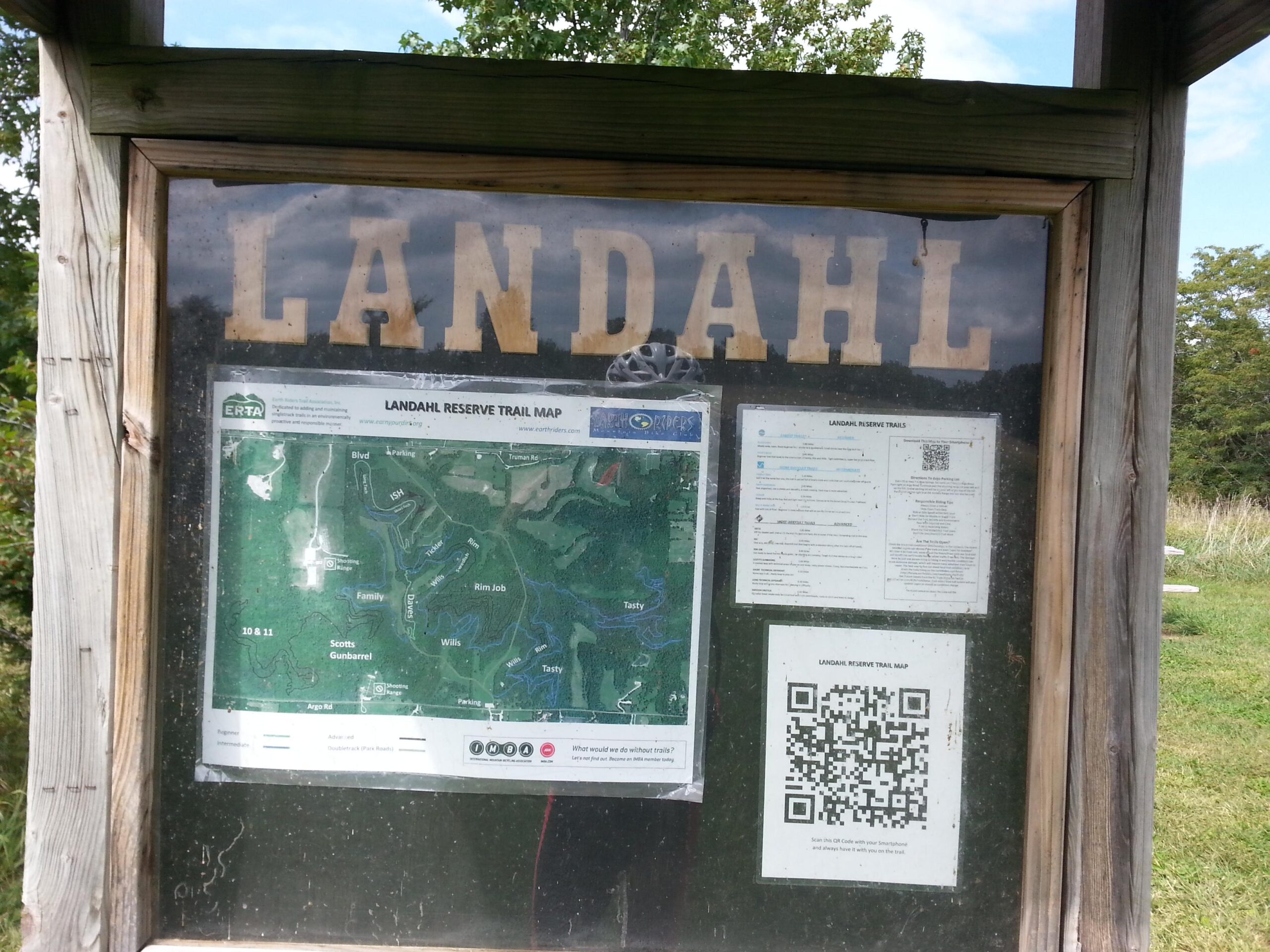 A wooden sign displaying the title "LANDAHL" at the top, with a trail map titled "LANDAHL RESERVE TRAIL MAP" underneath. The map outlines various trails in the reserve, while instructions and a QR code for more information are provided on the side. The background features trees and a grassy area, indicating an outdoor setting. Landahl Park Reserve mountain bike trail.