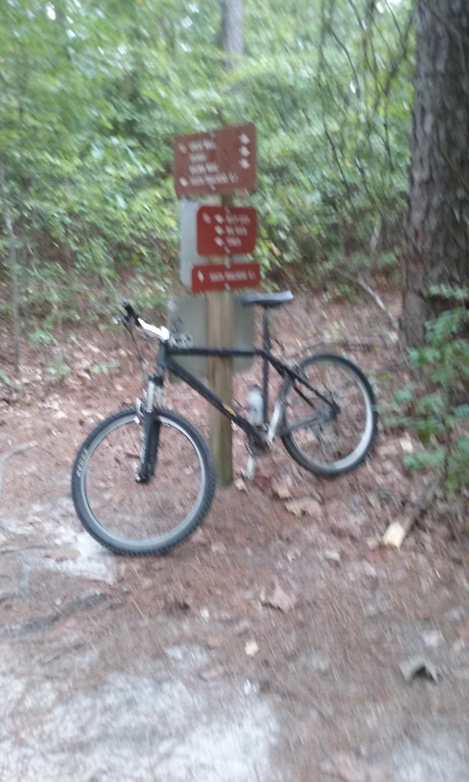 A black mountain bike leaning against a wooden trail sign surrounded by trees and fallen leaves. The sign displays various trail directions and distances in a forested area. Great Wall Loop mountain bike trail.