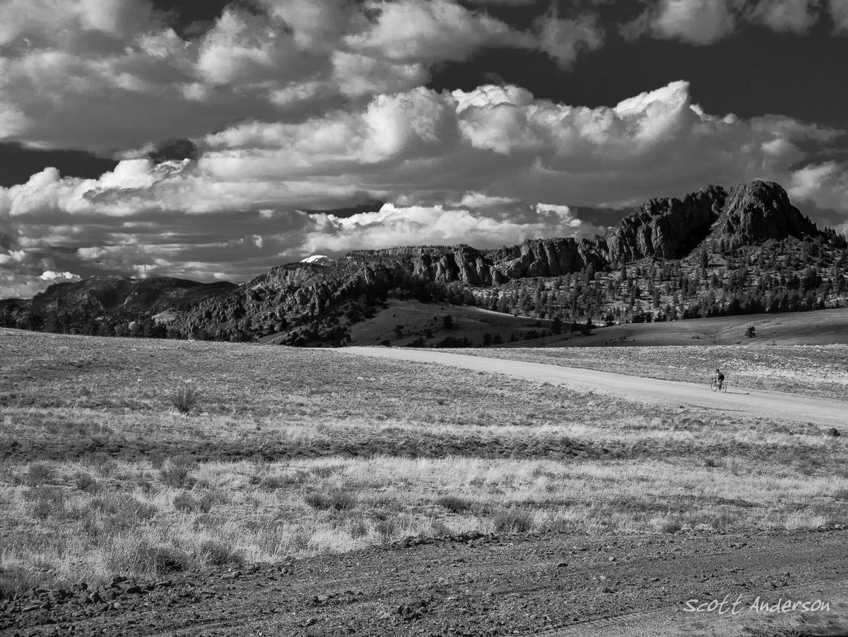 A black and white landscape image featuring a wide open field in the foreground with sparse vegetation, leading to a winding dirt road. In the background, rugged mountains rise against a dramatic sky filled with clouds. A cyclist can be seen riding along the road, adding a sense of scale to the expansive natural scenery. Road #187 mountain bike trail.