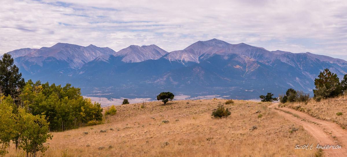 Panoramic view of a mountainous landscape featuring a range of peaks under a cloudy sky. In the foreground, grassy plains and sparse trees create a serene natural setting, while a dirt road winds through the scene. Road #185.D mountain bike trail.