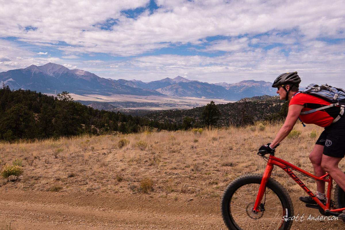 A cyclist riding a mountain bike on a dirt trail, surrounded by a scenic view of mountains and valleys under a partly cloudy sky. Trail #1434 mountain bike trail.