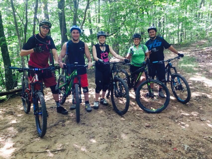 A group of five mountain bikers, wearing helmets and biking gear, stand together on a dirt trail surrounded by greenery. They each hold their bikes, smiling and posing for the photo. Bent Creek mountain bike trail.