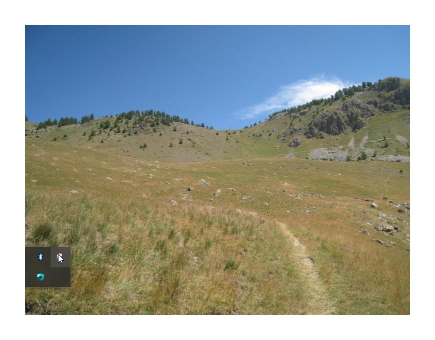 A scenic view of a grassy hillside with a clear blue sky, dotted with a few clouds. The landscape features gentle slopes, patches of trees on the hills, and a winding dirt path leading into the distance. The foreground shows expansive grassland with scattered rocks. Col de Tramouillon mountain bike trail.