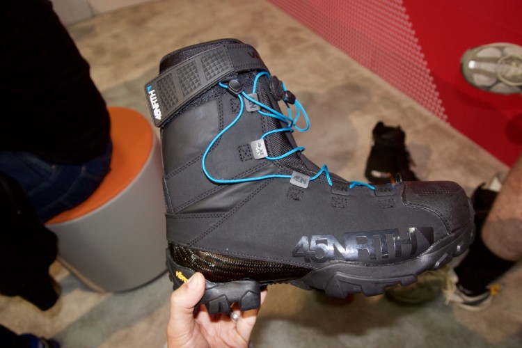 A black winter boot with blue laces and a textured upper, featuring a carbon fiber-look section on the sole, labeled "45NRTH." The boot is held in a hand against a neutral background, with a person seated in the background.