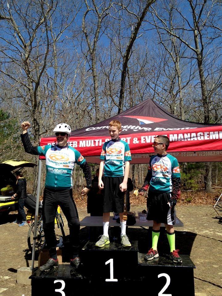 Three young cyclists celebrate their achievements on a podium at a biking event, with the first-place winner raising their fist in victory. They are wearing matching jerseys, and a tent in the background displays the words "Competitive Events Management." The scene is set in a wooded area with bare trees, indicating early spring.