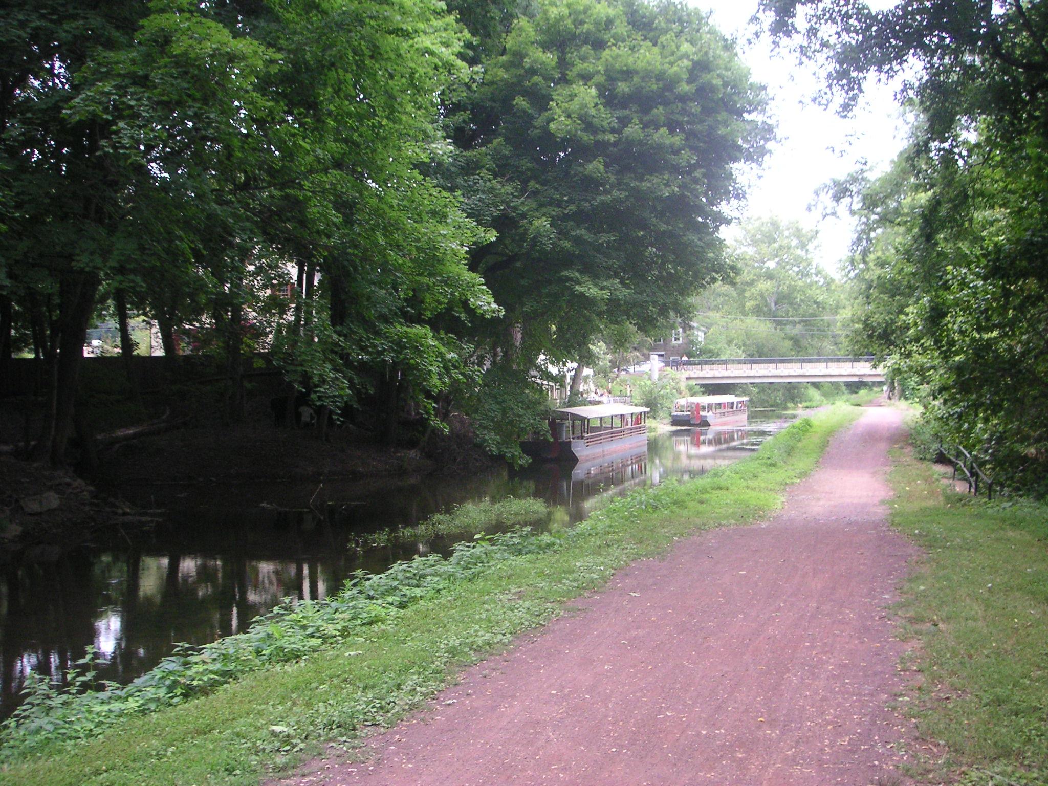 A serene pathway lined with greenery beside a calm canal, featuring two boats docked on the water. A bridge spans the canal in the background, surrounded by lush trees and foliage. The scene conveys a peaceful, natural setting suitable for walking or cycling. Bridge To Bridge - D&r Canal mountain bike trail.