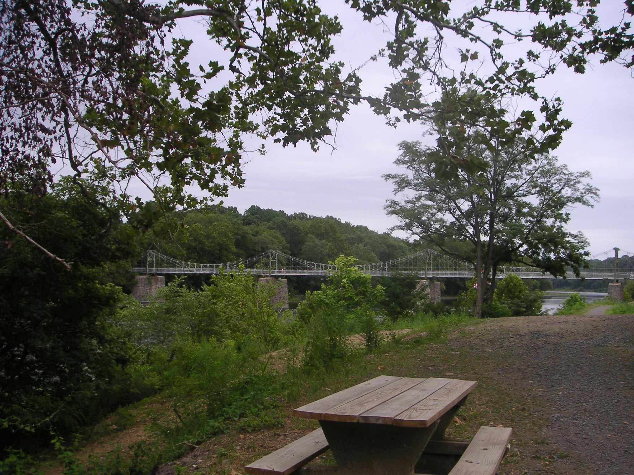 A scenic view of a suspension bridge spanning a river, surrounded by lush green trees and foliage. In the foreground, there is a picnic table made of wood and concrete, set against a gravel path leading to the water. The sky is overcast, adding a serene atmosphere to the landscape. Bridge To Bridge - D&r Canal mountain bike trail.