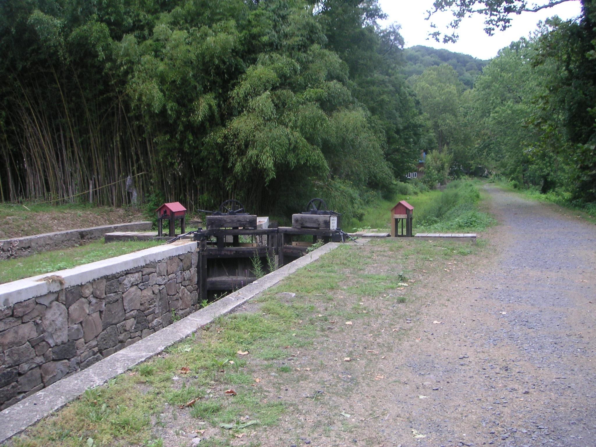 A scenic view of a canal lock system surrounded by greenery. The image features a stone wall structure with wooden components, including a wheel, set in a grassy area. In the foreground, a gravel path leads towards the lock, with lush trees and bamboo in the background, creating a tranquil, natural setting. Bridge To Bridge - D&r Canal mountain bike trail.