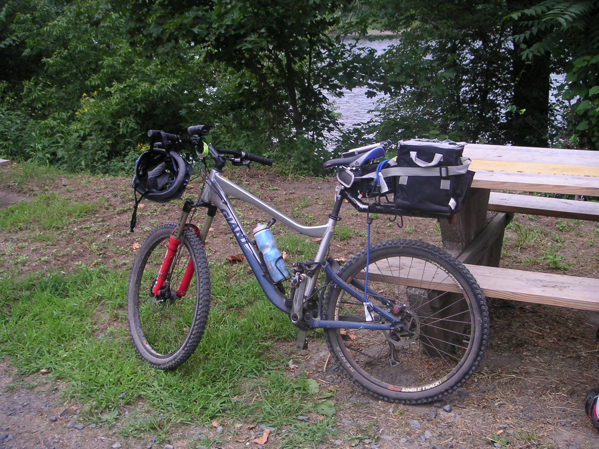 A silver mountain bike parked on grassy ground near a river, with a black helmet hanging from the handlebars and a water bottle attached. A picnic table is visible in the background, surrounded by greenery. Bridge To Bridge - D&r Canal mountain bike trail.