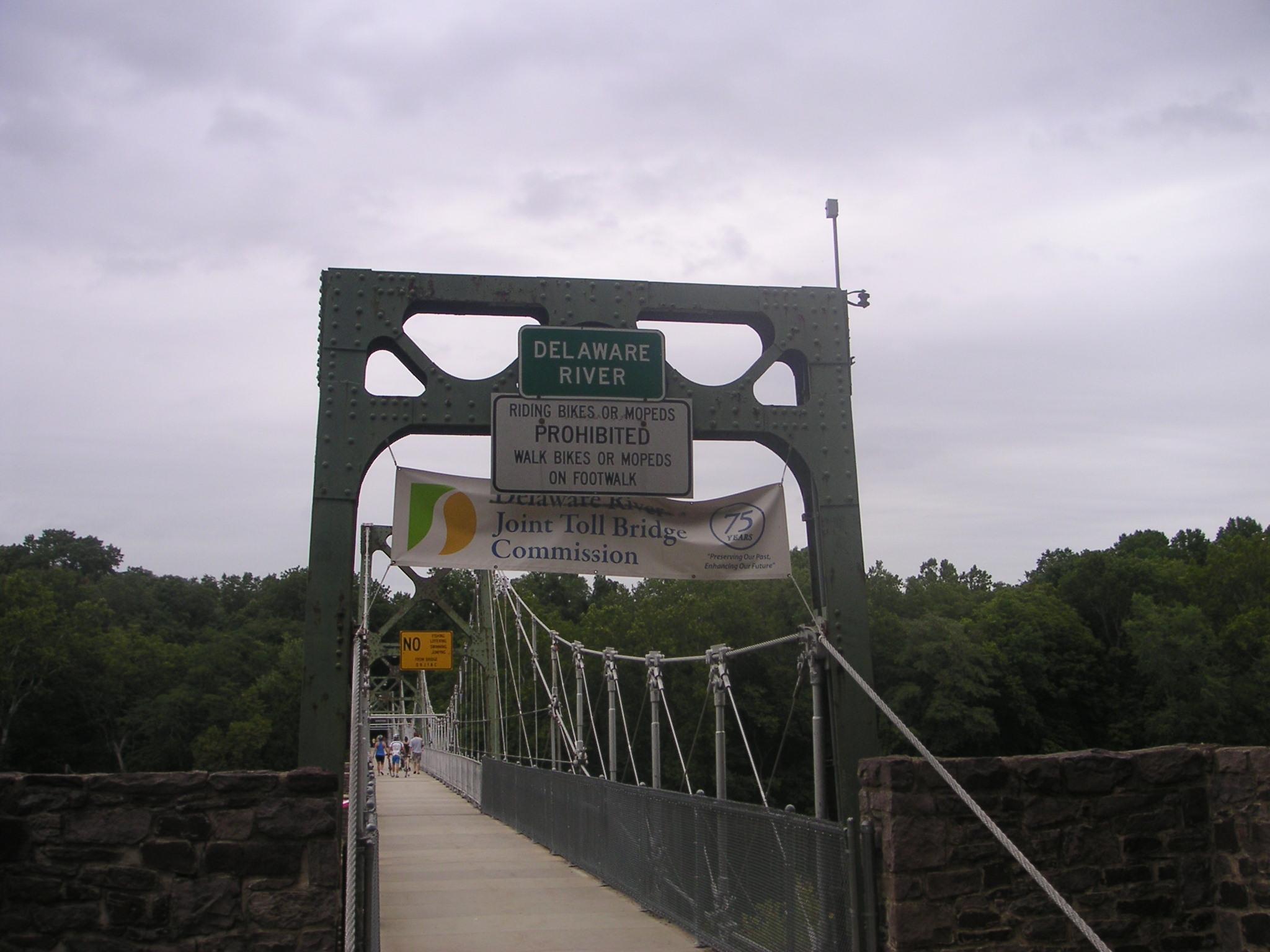 A suspension bridge over the Delaware River, featuring a metal frame and a walkway. Visible signs indicate restrictions on biking and riding scooters. A banner for the Delaware River Joint Toll Bridge Commission celebrates 75 years, with lush greenery in the background under a cloudy sky. Bridge To Bridge - D&r Canal mountain bike trail.
