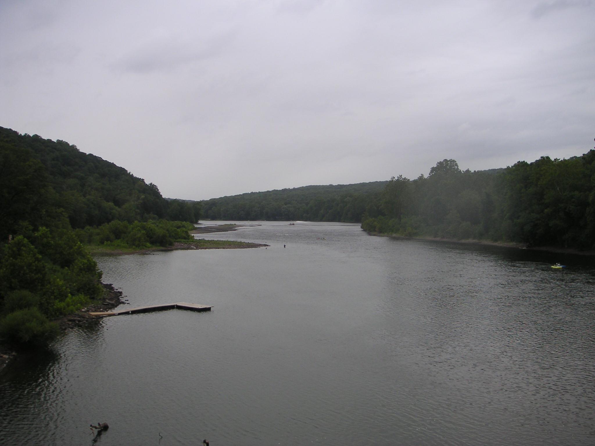 A scenic view of a river winding through lush greenery under a cloudy sky. A wooden dock extends into the water on the left side, while trees line both shores. The river reflects the overcast sky, creating a tranquil atmosphere. Bridge To Bridge - D&r Canal mountain bike trail.