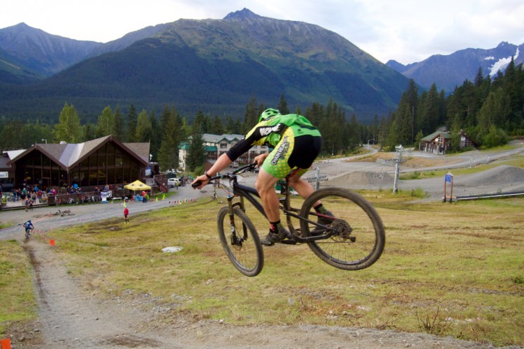 A mountain biker in mid-air jumps off a dirt trail in a scenic outdoor setting, surrounded by mountains and trees. In the background, a building with large windows is visible, along with other cyclists and spectators. The biker is wearing a green helmet and a jersey with a green and black design.