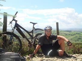 Specialized Enduro Pro: A person sitting on the ground beside a mountain bike, with a scenic landscape of rolling hills and a blue sky in the background. The person is wearing a helmet and cycling gear, enjoying a moment of rest after biking.