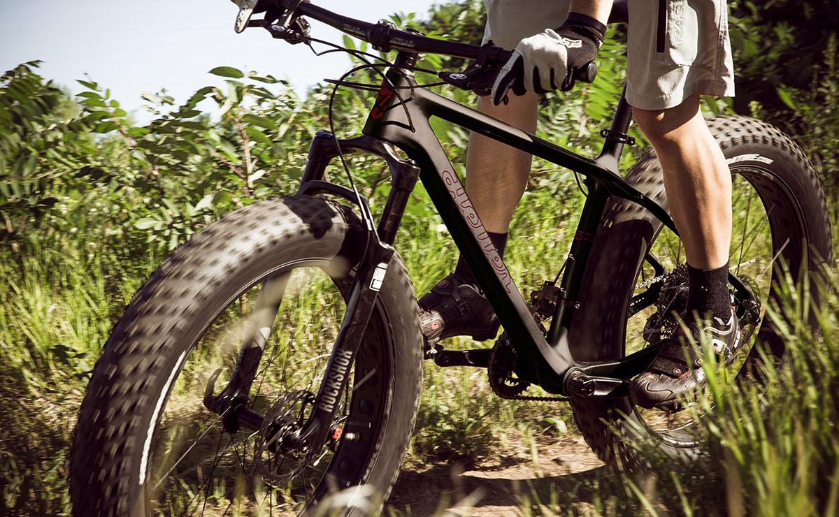 Heller Bloodhound: A close-up view of a person's legs riding a fat-tire mountain bike on a grassy trail, with lush greenery in the background. The cyclist is wearing gloves and a light-colored outfit, emphasizing the rugged outdoor environment.