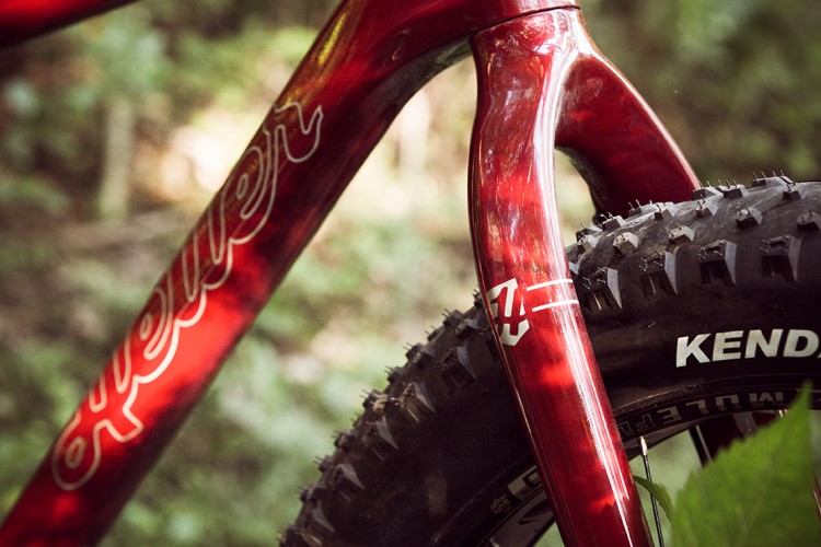 A close-up image of a red bicycle frame with the brand name partially visible. The foreground features a thick tire with the brand "Kenda" printed on it. The background shows a blurred green natural setting, indicating an outdoor environment.
