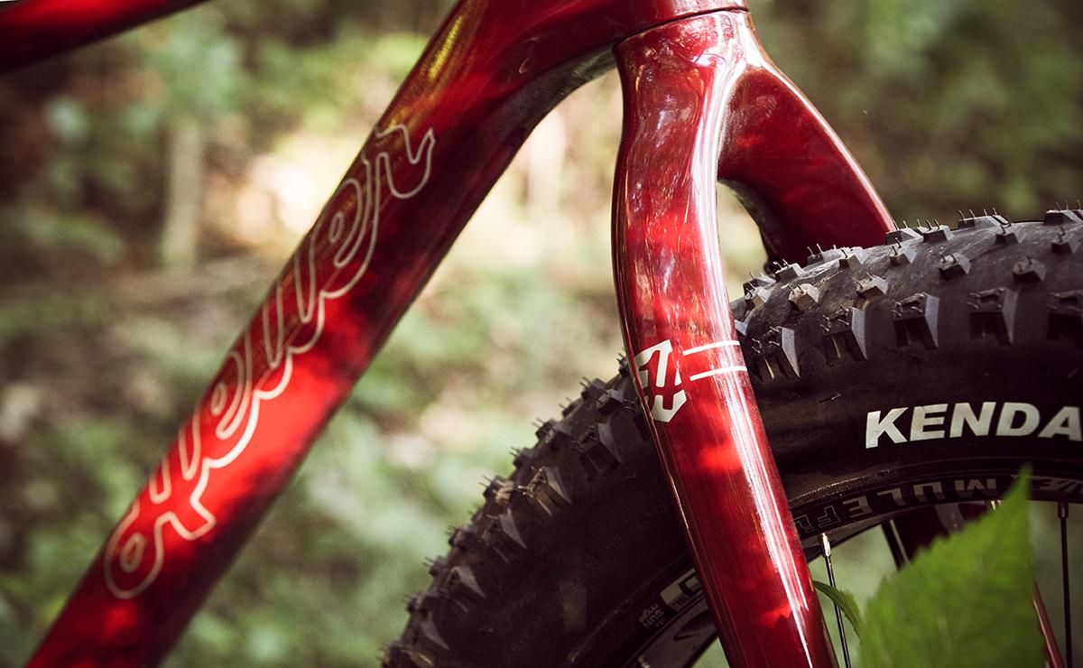 Heller Bloodhound: Close-up of a red bicycle frame with a visible logo and a thick, knobby tire labeled "Kenda," set against a blurred forest background. The image emphasizes the bike's sturdy design and rugged features.