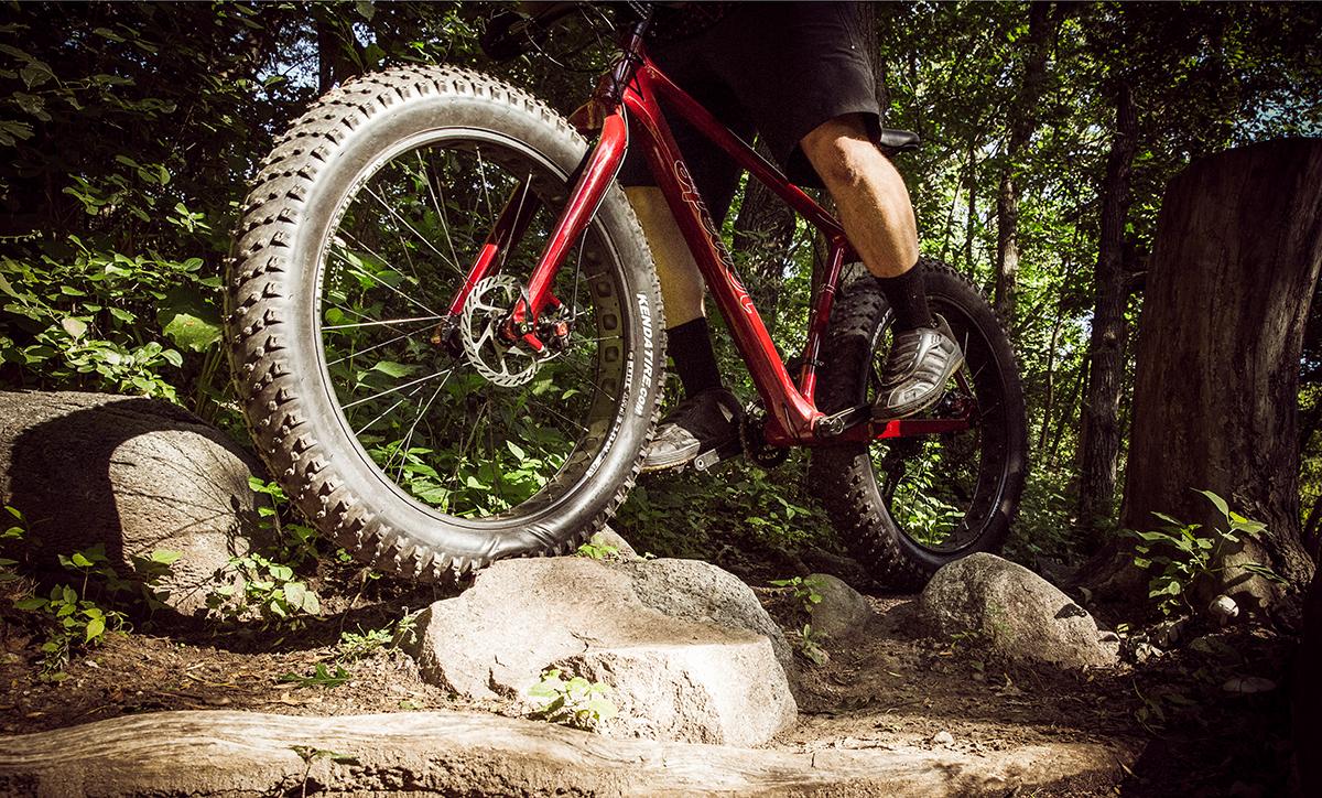 Heller Bloodhound: A close-up view of a mountain biker riding over rocky terrain on a fat bike. The emphasis is on the large, knobby tire and the biker's foot positioned on the pedal, with a lush green background of trees and foliage. Sunlight filters through the trees, highlighting the bike's red frame.